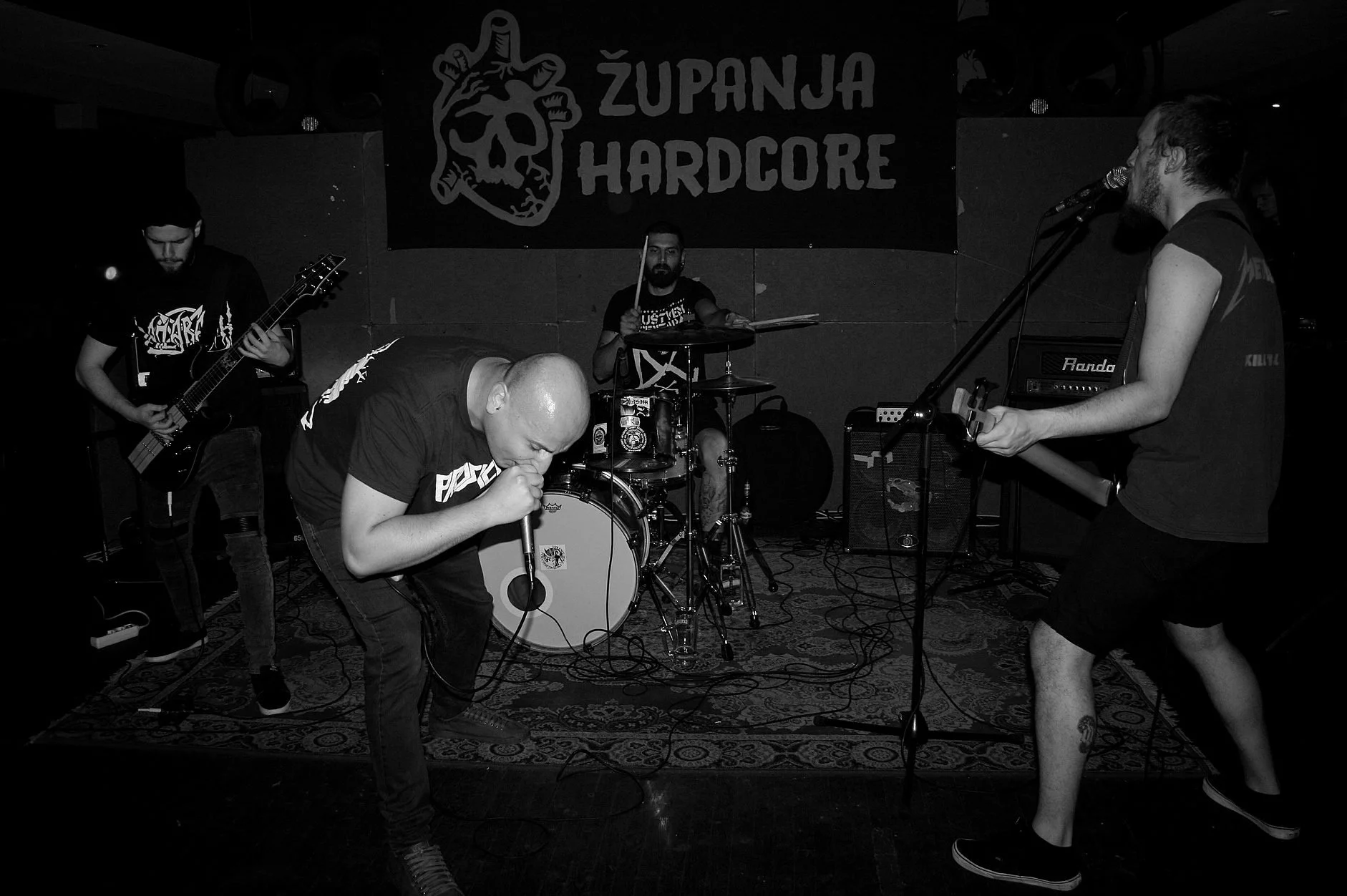 Black and white photo of a live hardcore music band performing on stage with a banner reading 'ŽUPANJA HARDCORE' in the background. The band members are playing various instruments, including electric guitar, drums, and vocals, with one member singin