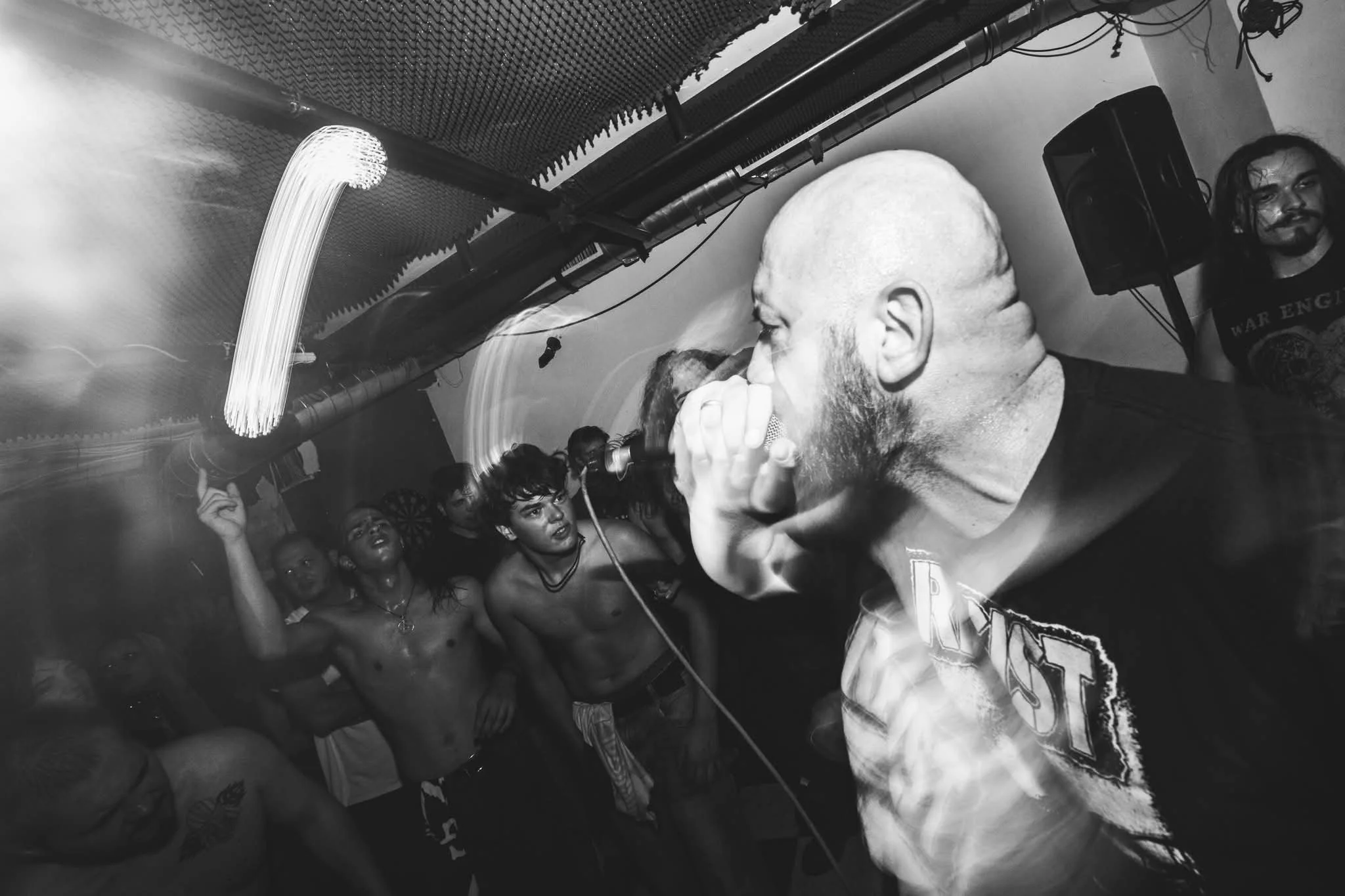 A black and white photo of a live concert. The audience is crowded and mostly shirtless. The singer, with a beard and short hair, is holding a microphone close to his mouth. There is a man with long hair and a mustache standing near a speaker on the 
