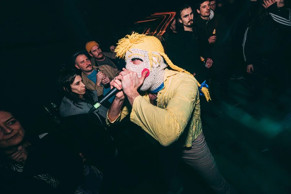 Person wearing a large, colorful, knitted mask with exaggerated features and yellow hair, singing into a microphone in a crowded nightclub.