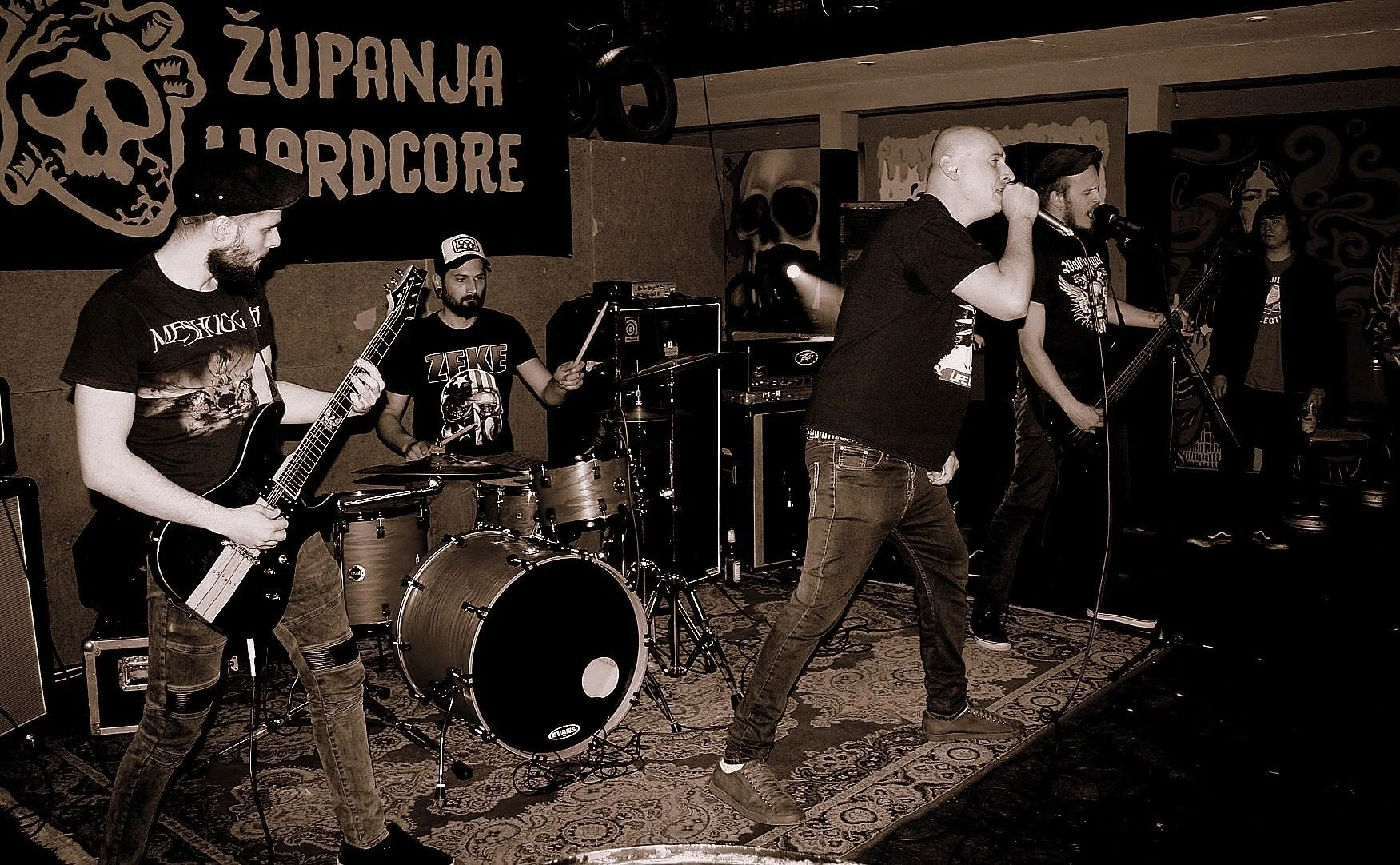 A band performing on stage with four members: a guitarist, drummer, and two vocalists. The backdrop features a banner with a skull and the words 'ZUPANJA HARDCORE'. The scene is indoors with sound equipment and a patterned rug.