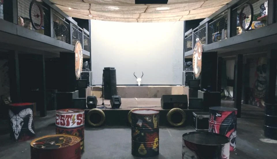 Indoor concert or event space with stage, speakers, and microphone, surrounded by graffiti-covered drums and artwork, with balcony or mezzanine level on both sides.