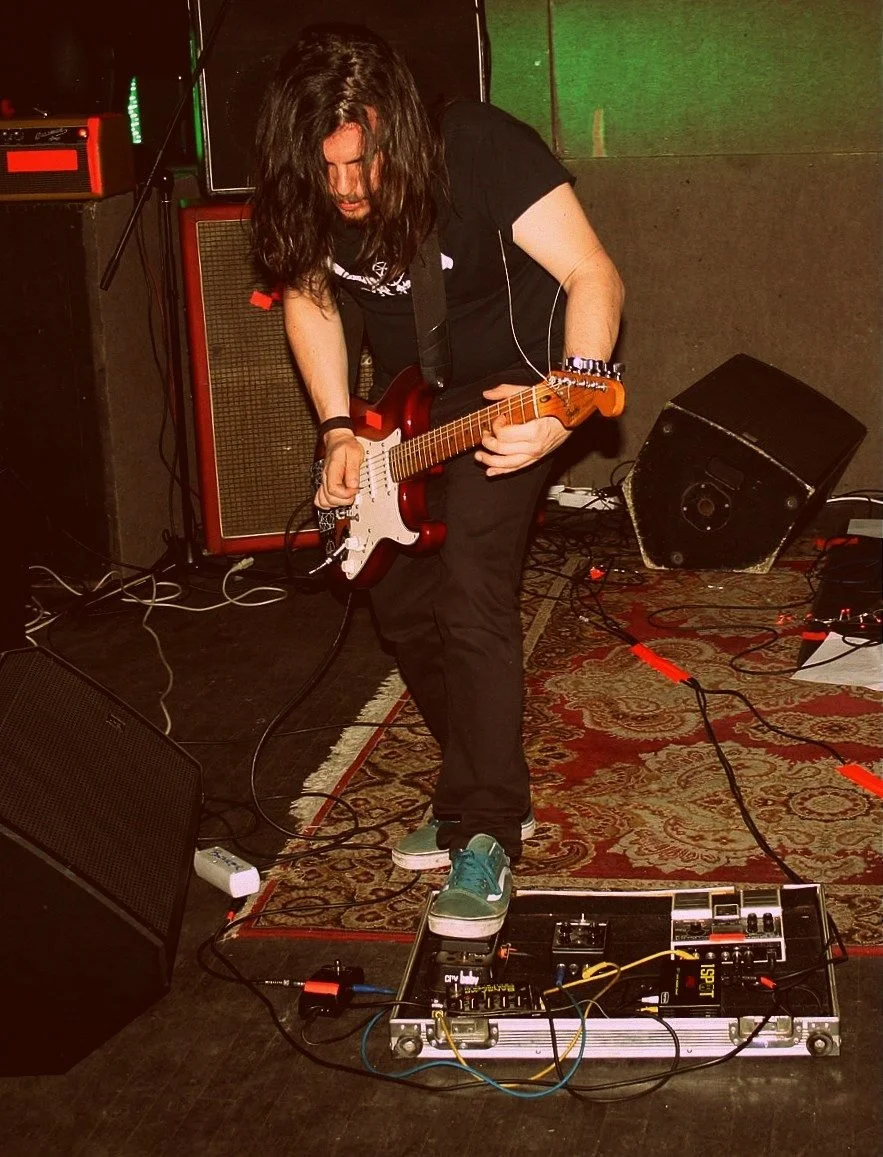 A person with long hair playing an electric guitar on stage, surrounded by amplifiers, pedals, and cables, wearing casual clothes and sneakers.