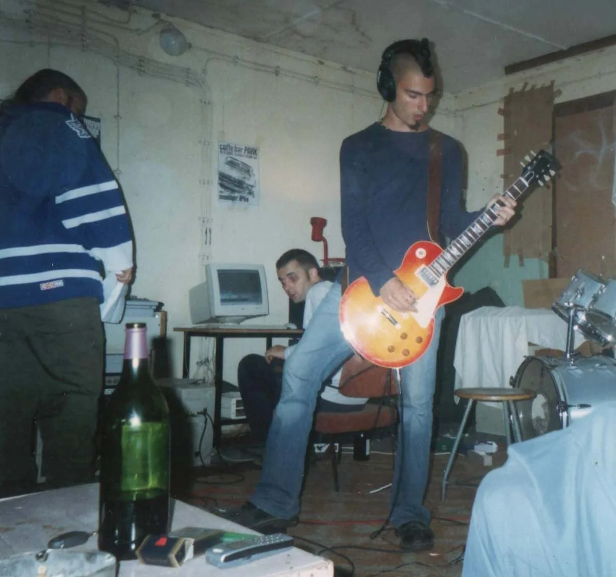 Three young men in a room; one playing an electric guitar, another seated at a computer, and the third standing beside with their head down. Items include a large bottle on a table, a remote control, and a drum set.