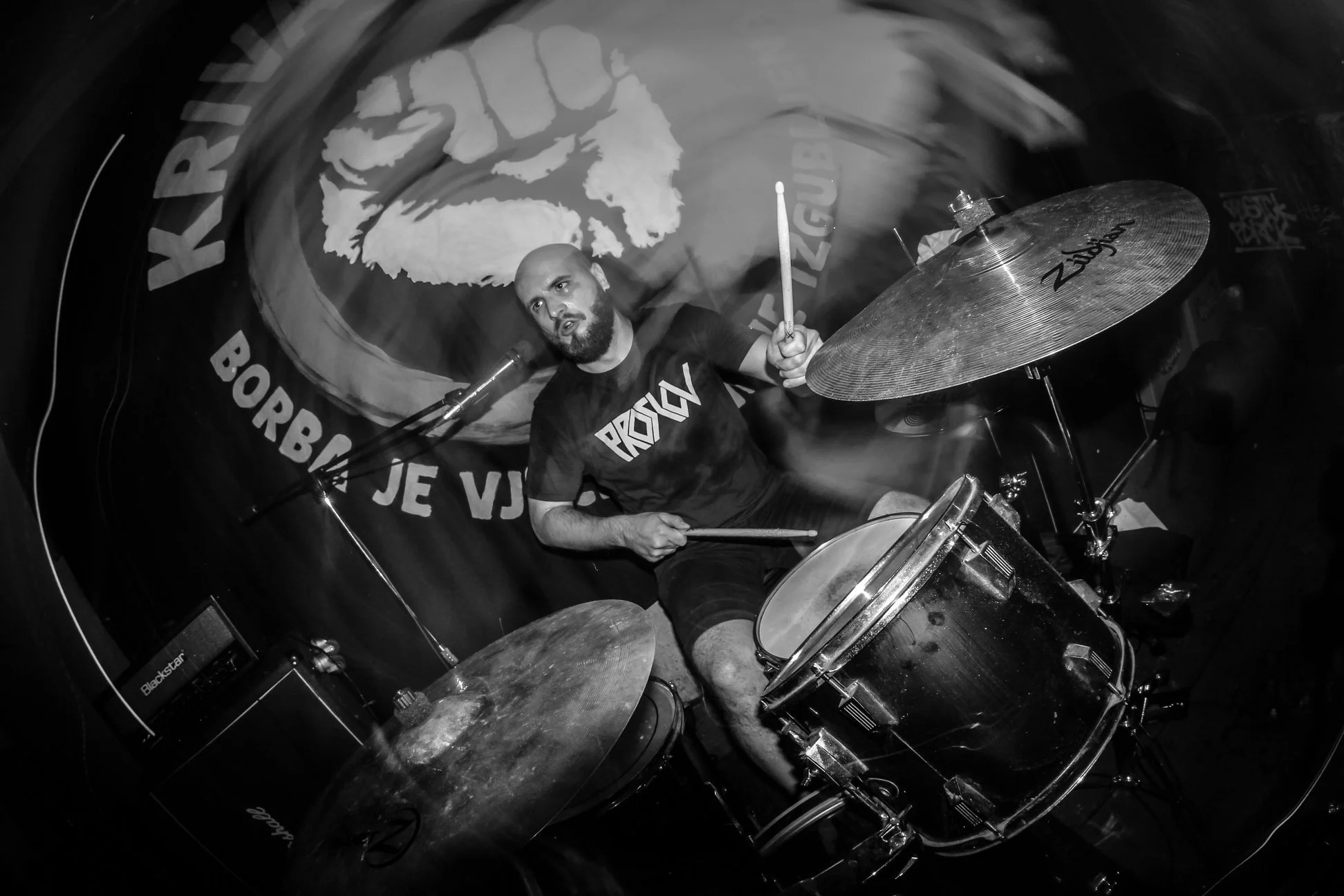 Black and white photo of a drummer playing drums on a stage. The background features a large circular logo with a fist and the word 'KRIVA ISTINA' visible. The drummer is wearing a T-shirt with the word 'PROSLOV' and has a beard and bald head.