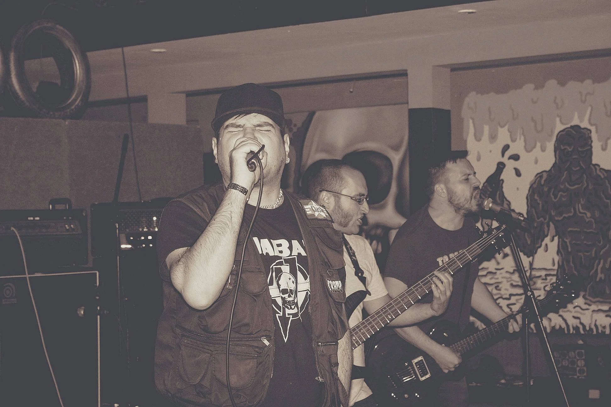 A band of four musicians performing on stage. The lead singer is at the front, holding a microphone with his eyes closed. Behind him, two others are playing guitars — one with a cigar in his mouth and the other with glasses. The scene is set in a dim
