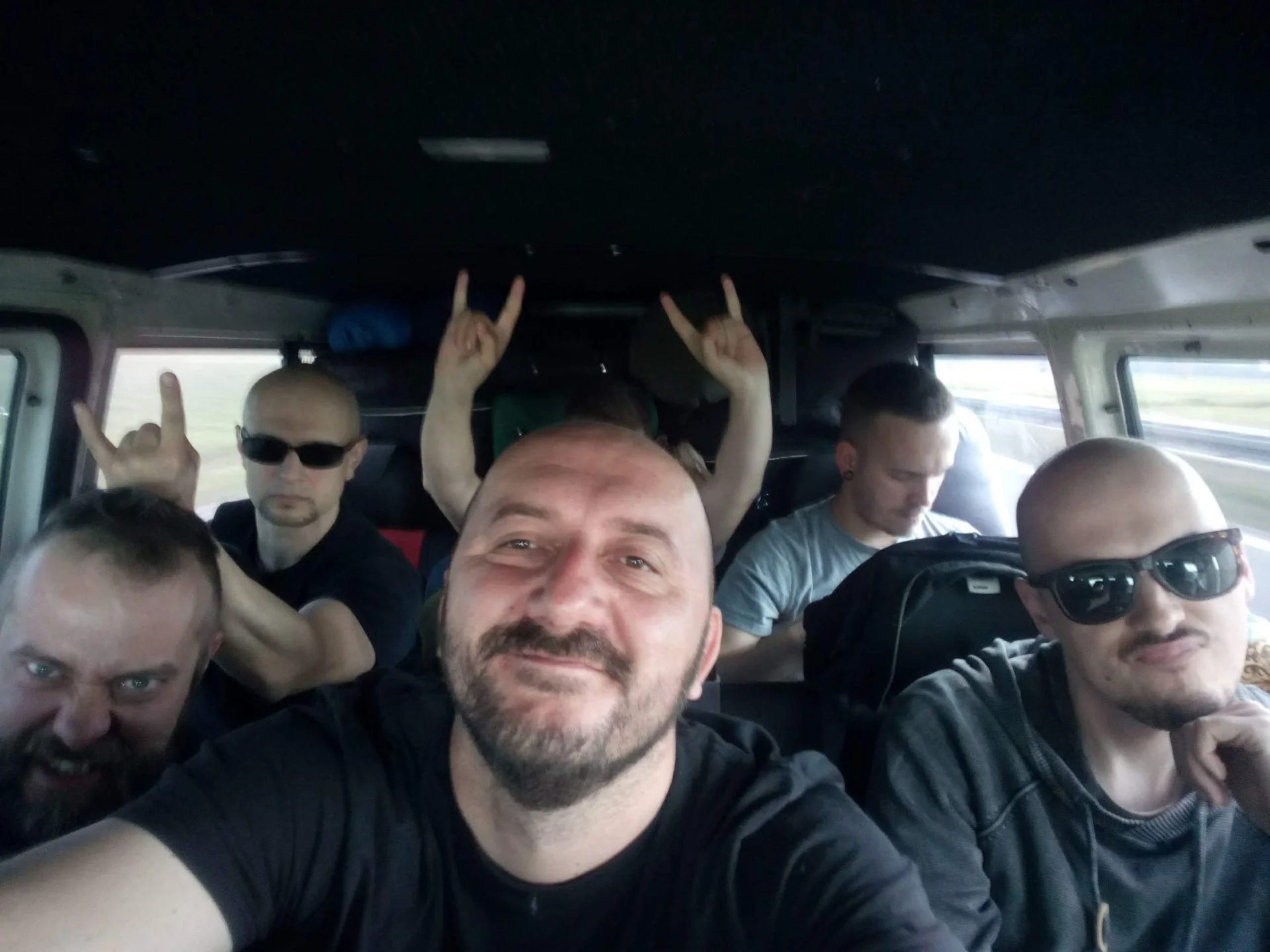 Group of people inside a vehicle, some making rock hand gestures, with sunglasses and casual clothing, smiling and posing for a selfie.