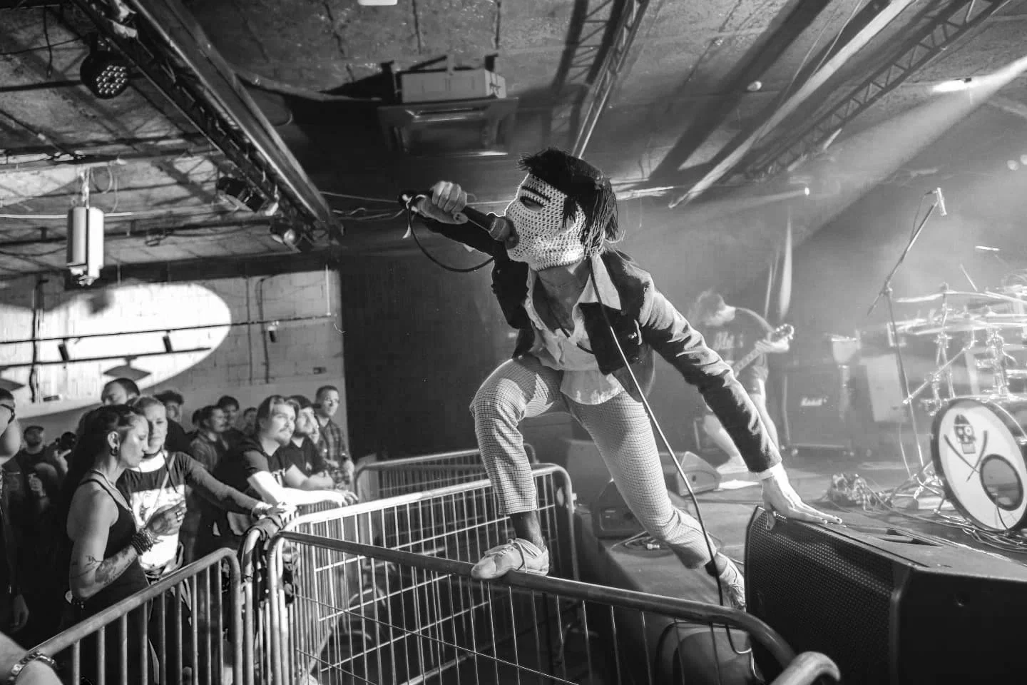 A live music performance at a concert venue. The lead singer is wearing a mask and casual clothing, singing into a microphone while crouching on the stage. The audience is close to the stage, watching intently, with some reaching out.
