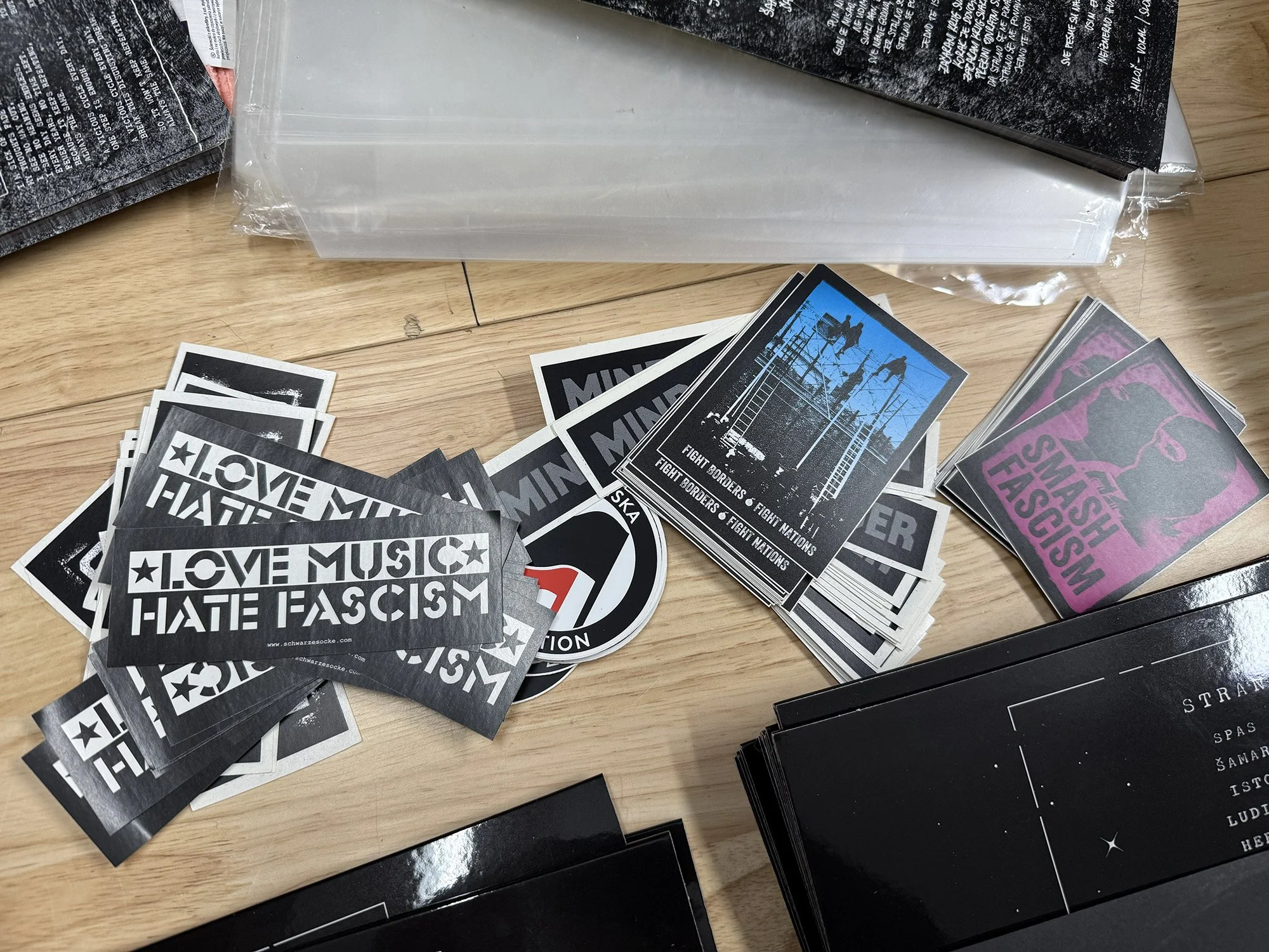 A collection of stickers with political, social, and protest messages spread out on a wooden surface. Some stickers include phrases like 'LOVE MUSIC HATE FASCISM' and 'FIGHT BORDERS' with images of barbed wire and people climbing fences.
