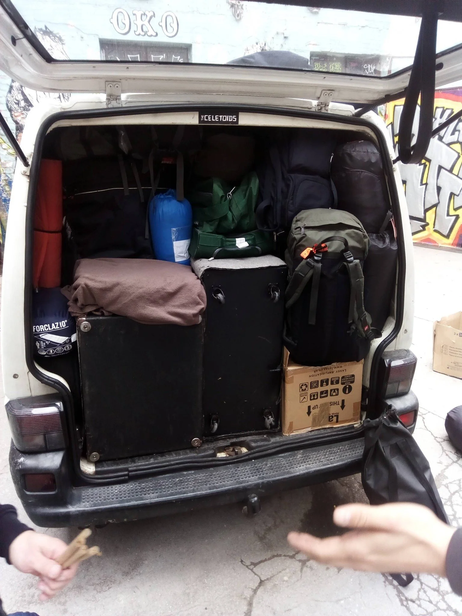 Back of a vehicle with an open hatch, filled with various bags, boxes, and gear. Two hands are visible at the bottom of the image, holding small objects.
