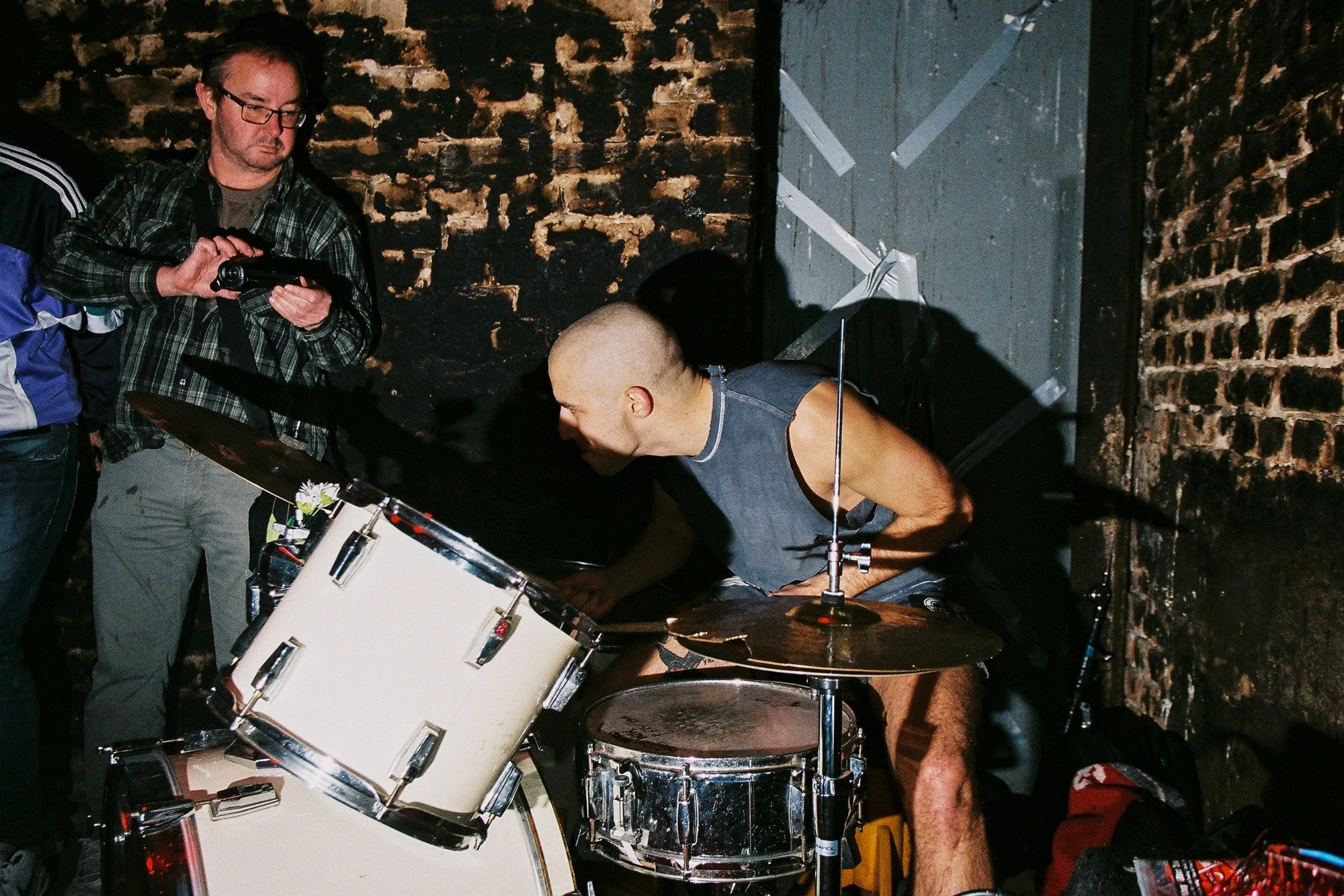 A drummer with a shaved head playing a white drum set with a dark background featuring a brick wall, while a man with glasses wearing a plaid shirt looks on, holding a camera or recording device.