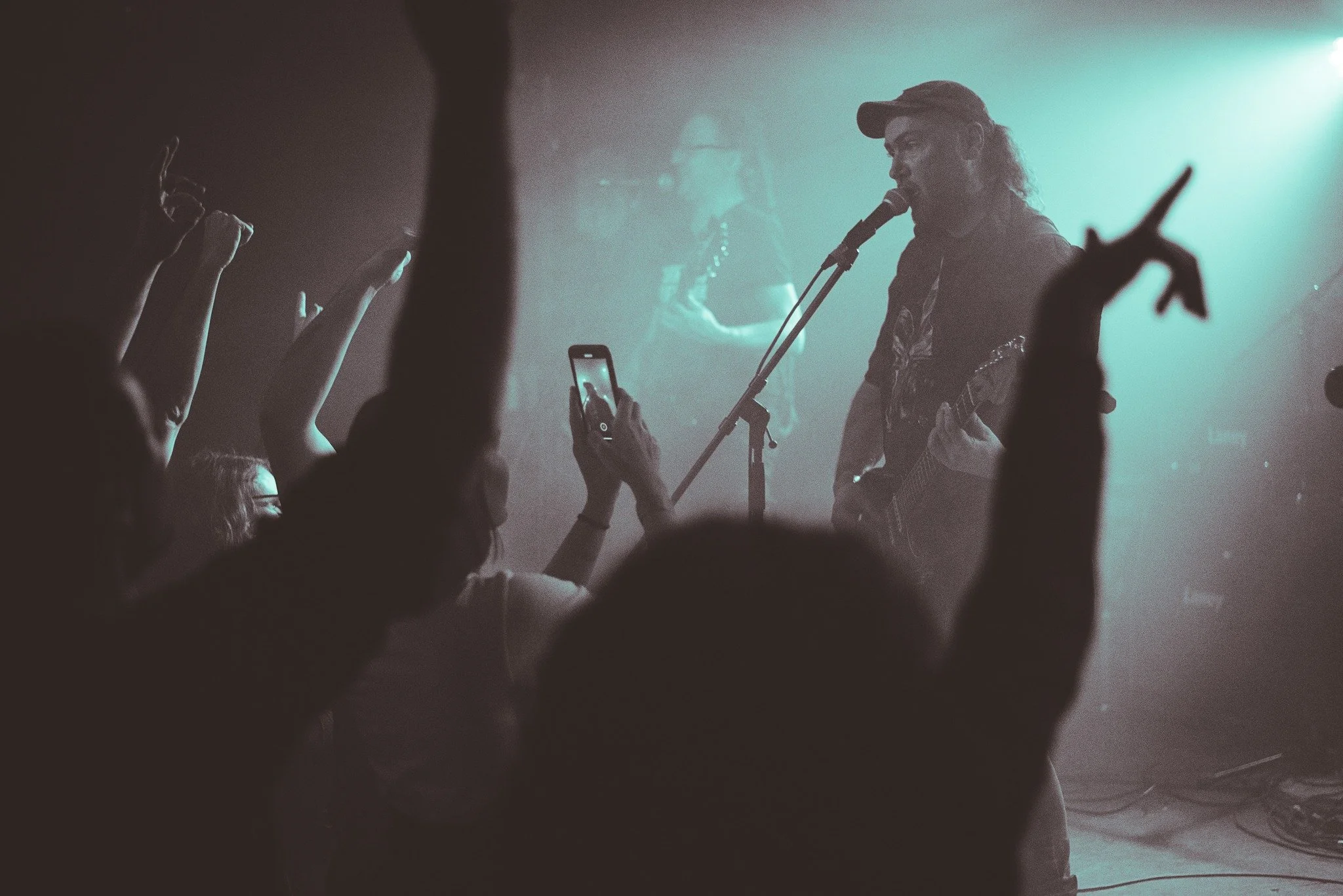 A musician with long hair, baseball cap, and guitar singing into a microphone on stage, with a crowd watching and reaching out, some holding phones capturing the performance. There's a blue-green haze from stage lighting.