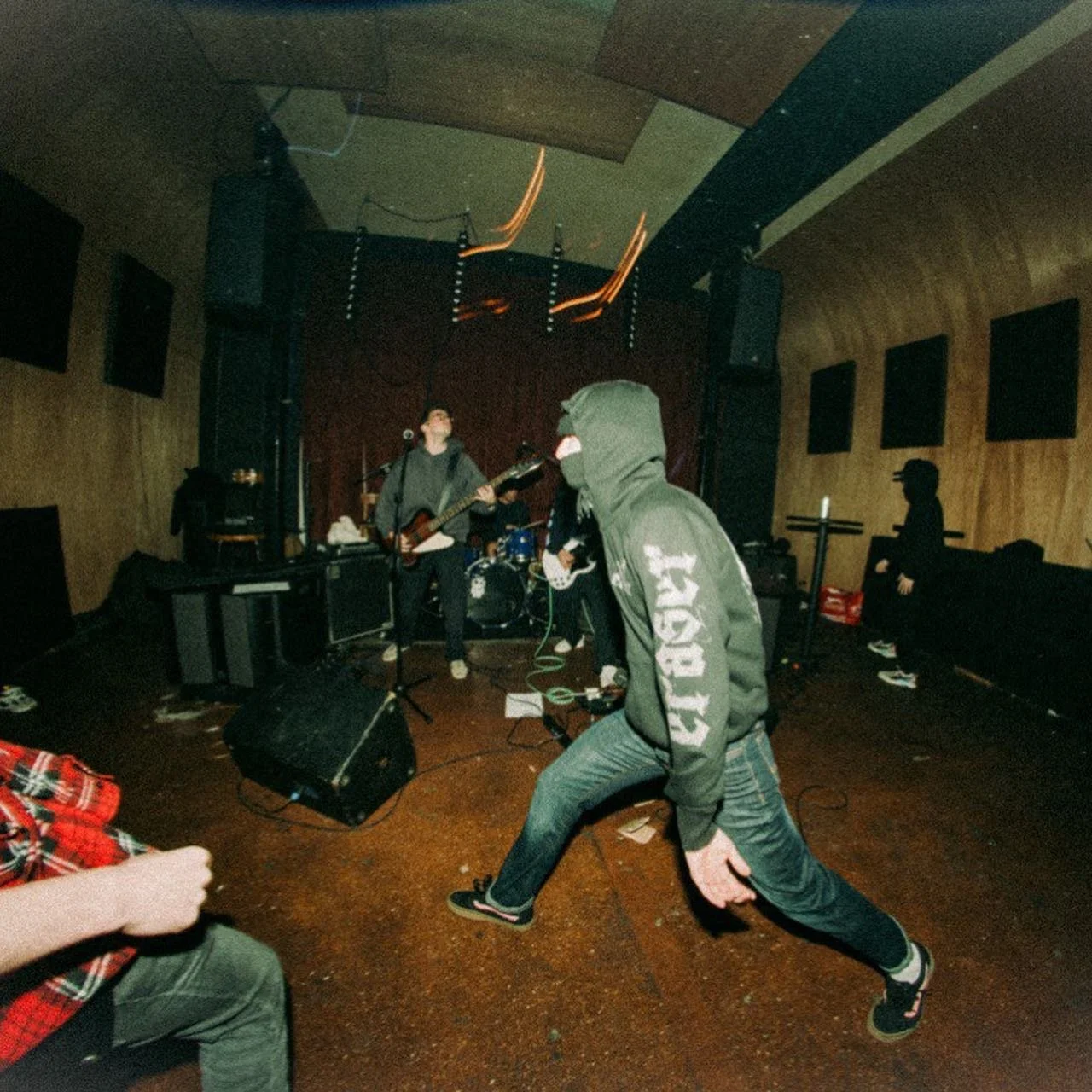 A band performing on stage with a person in the foreground dancing or moshing, wearing a hooded jacket with the words 'SAINT' on the sleeve, in a small music venue with wood-paneled walls and sound panels.