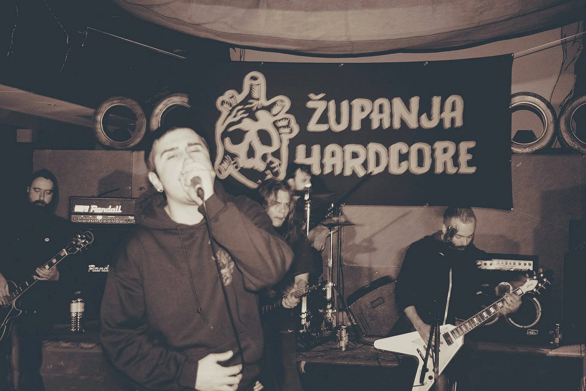 A band performing live on stage with a singer and musicians playing guitars and drums. A banner in the background reads 'ZUPANA HARDCORE' with a skull and snake logo.