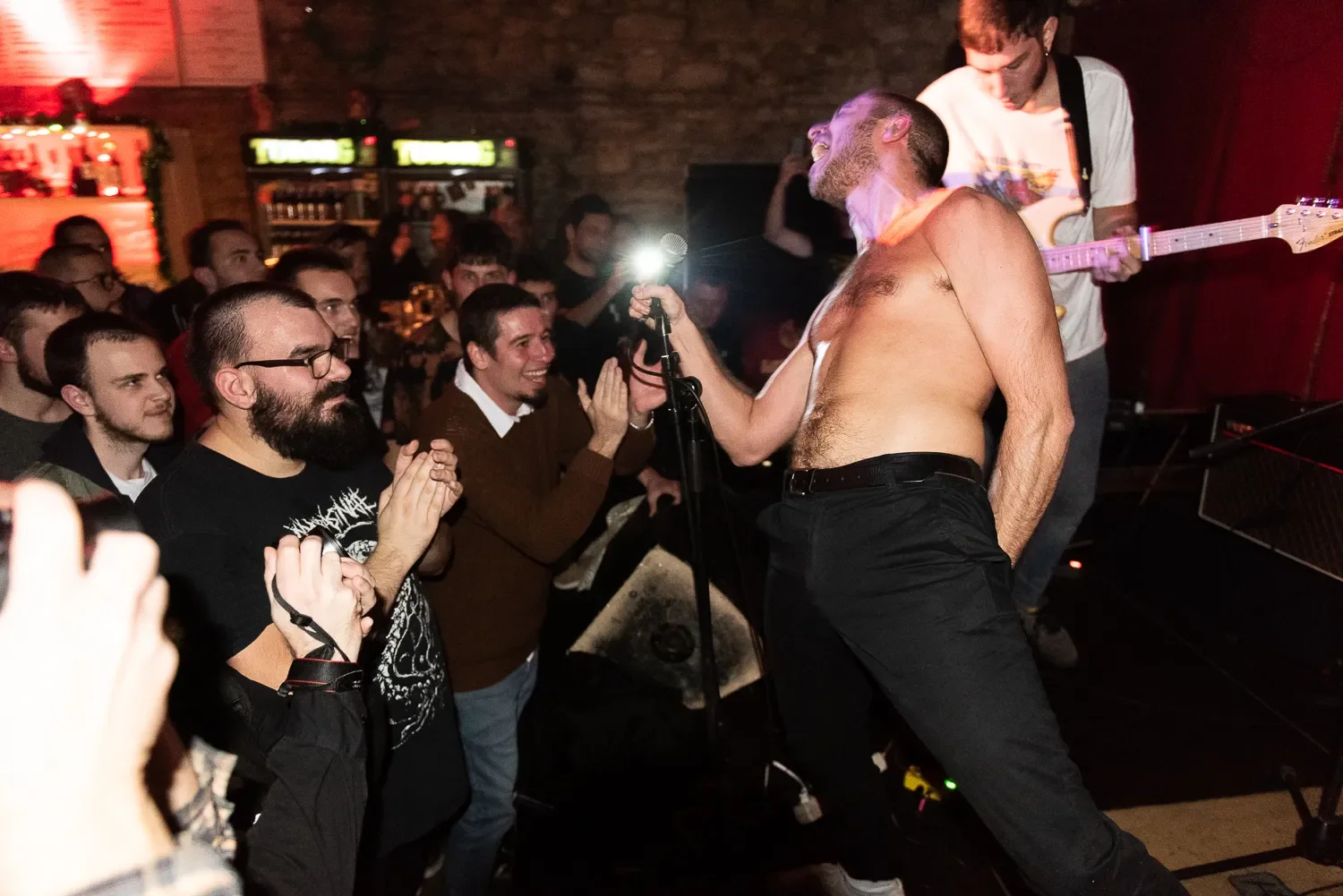 A shirtless man with a beard singing into a microphone on stage in front of an audience at a concert, with a guitarist in the background.