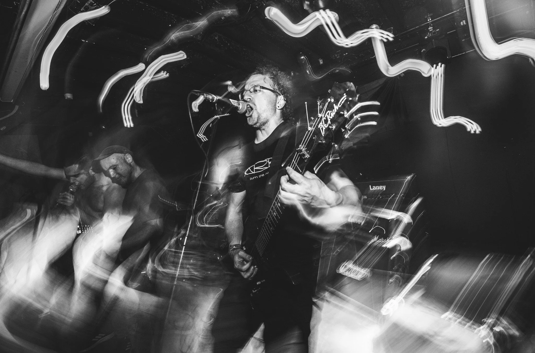 Black and white photo of a live music performance, featuring a singer with glasses passionately singing into a microphone and playing an electric guitar. Two other band members are visible, one with a microphone and the other focused on his instrumen