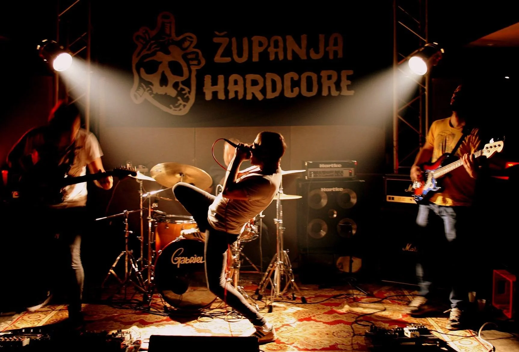 A live band performs on stage under bright spotlights at a concert titled 'Županja Hardcore', with two guitarists on either side, a drummer in the background, and a vocalist holding a microphone.