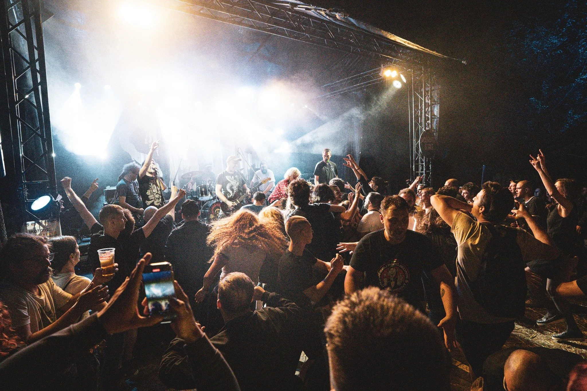 A live music concert with a band performing on stage at night. The crowd is dancing and enjoying the music, with some people holding drinks and others taking photos.