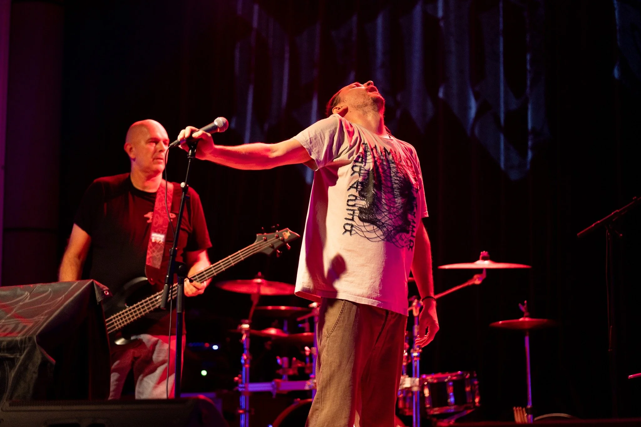 A band performing on stage with one singer holding a microphone, singing with his head tilted back. A guitarist is visible to the left, and drums are in the background. The stage is lit with warm, reddish lighting.