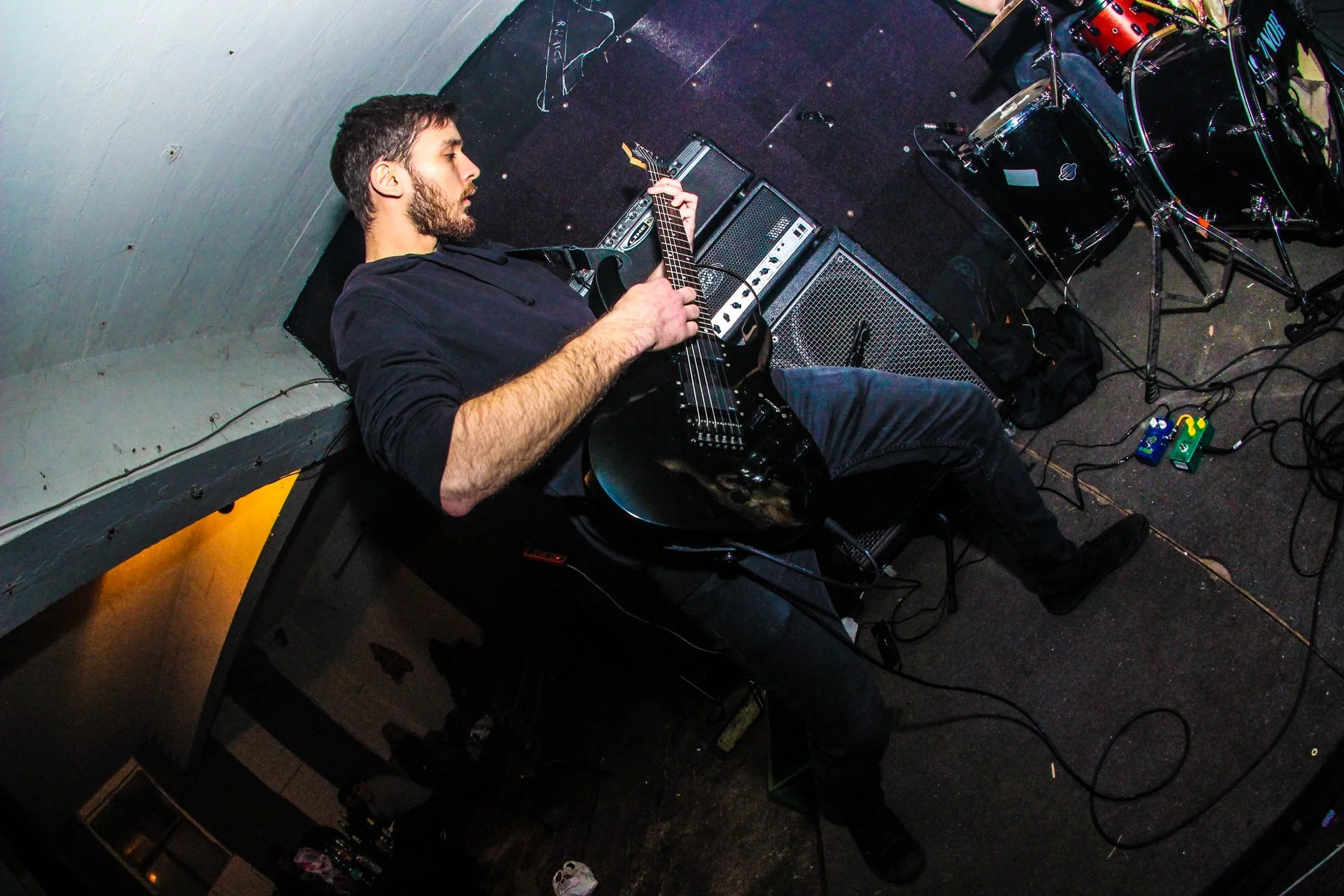 A man with a beard and dark hair playing an electric guitar on stage, surrounded by amplifiers and drum set, with a black floor and gray wall.