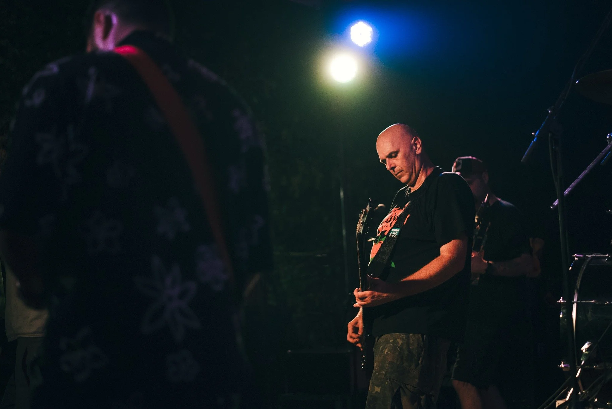 A band performing live on stage in a dark setting, with two musicians visible; one playing an electric guitar with his eyes closed, and the other partially visible, wearing camouflage pants.