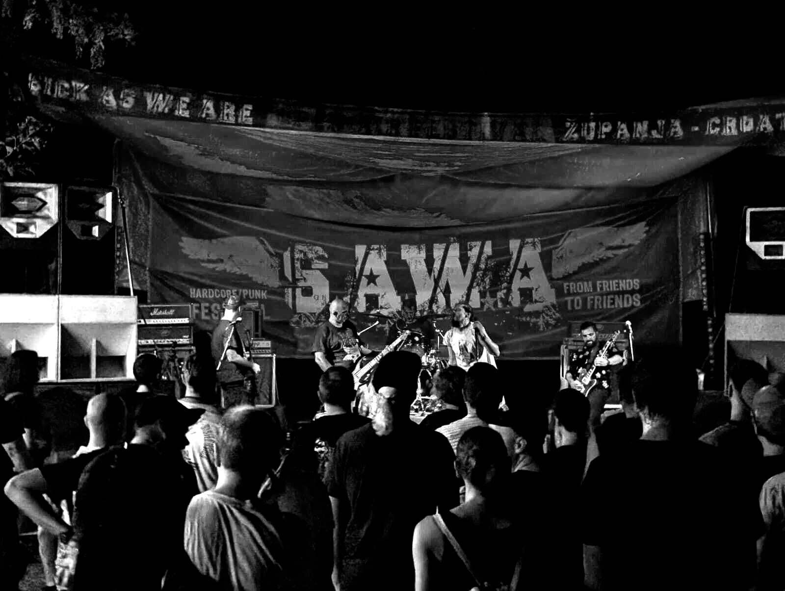 A live music concert with a band performing on stage, audience watching, stage decorated with a banner reading 'SAWAS' and 'from friends to friends', speakers on the sides.