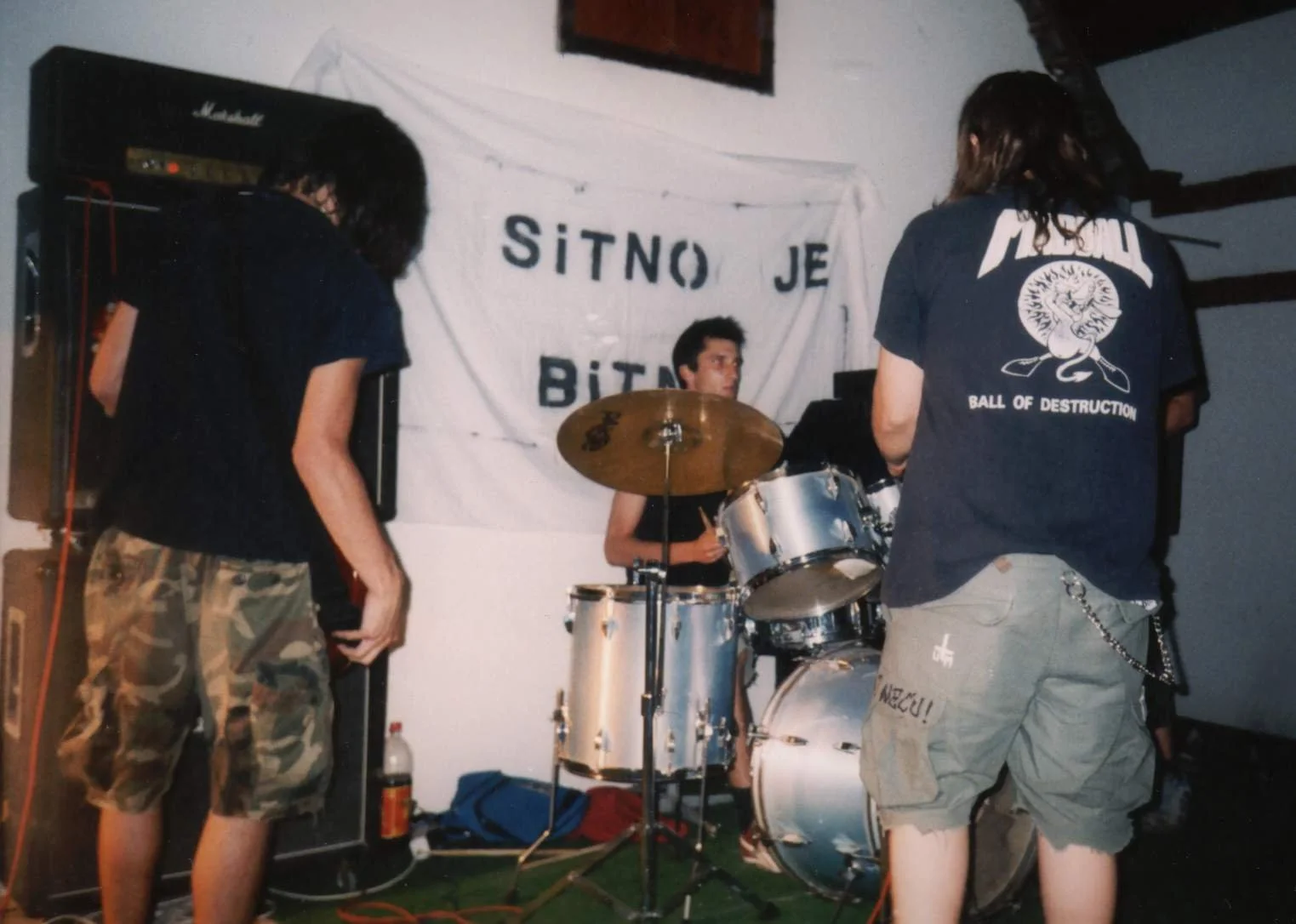 A band performing at a concert, with a drummer in the center and two guitarists on either side, one facing away and the other with their back to the camera, in a room decorated with a banner that reads 'SITNO JE BITNO'.