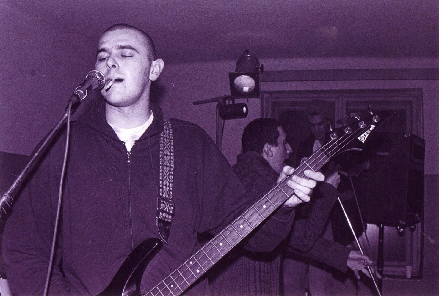 Young man with a shaved head singing into a microphone while playing an electric bass guitar, with two other men in the background in a dimly lit room.