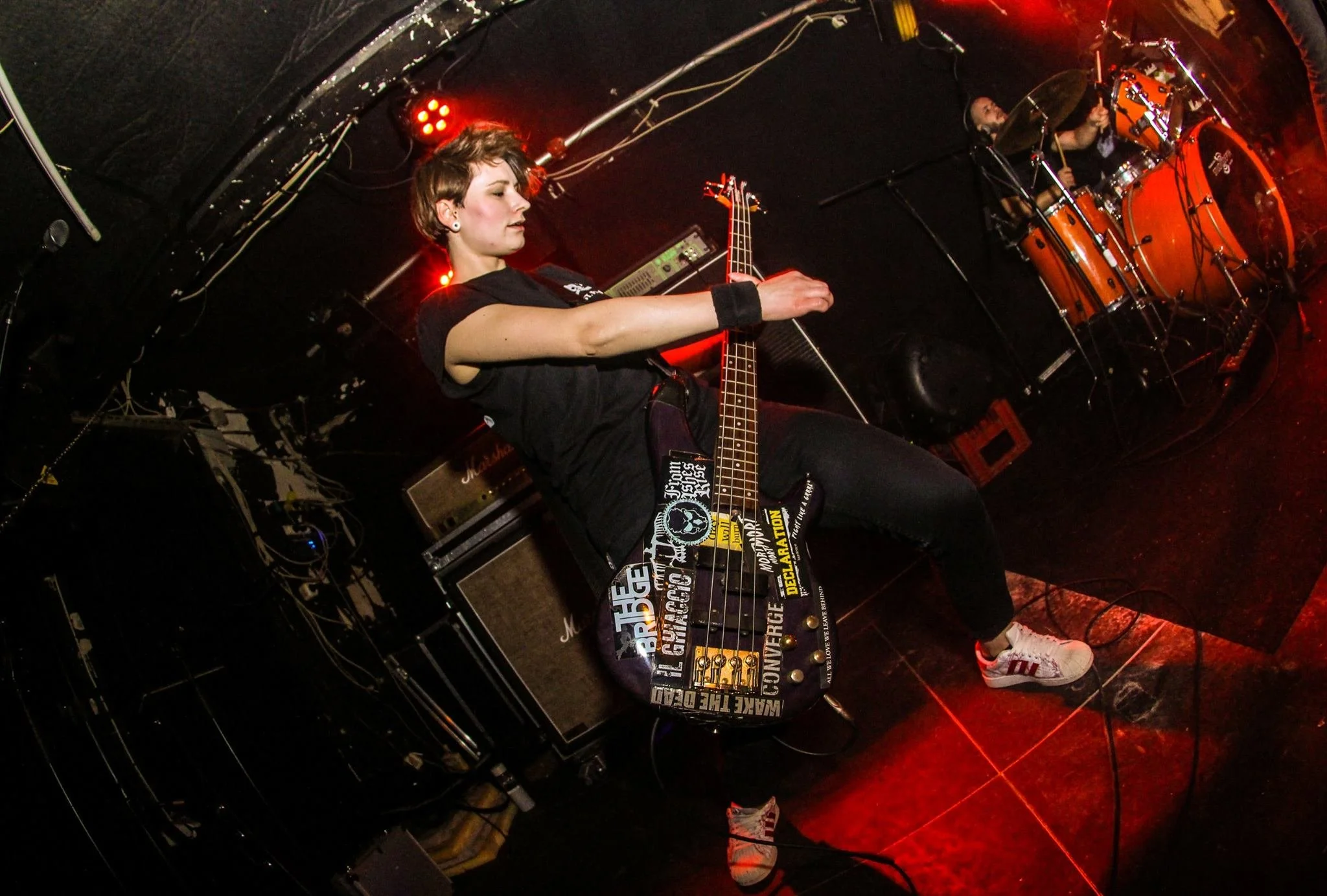 A female musician with short hair playing an electric bass guitar on stage, surrounded by music equipment and red stage lighting, with a drummer visible in the background.