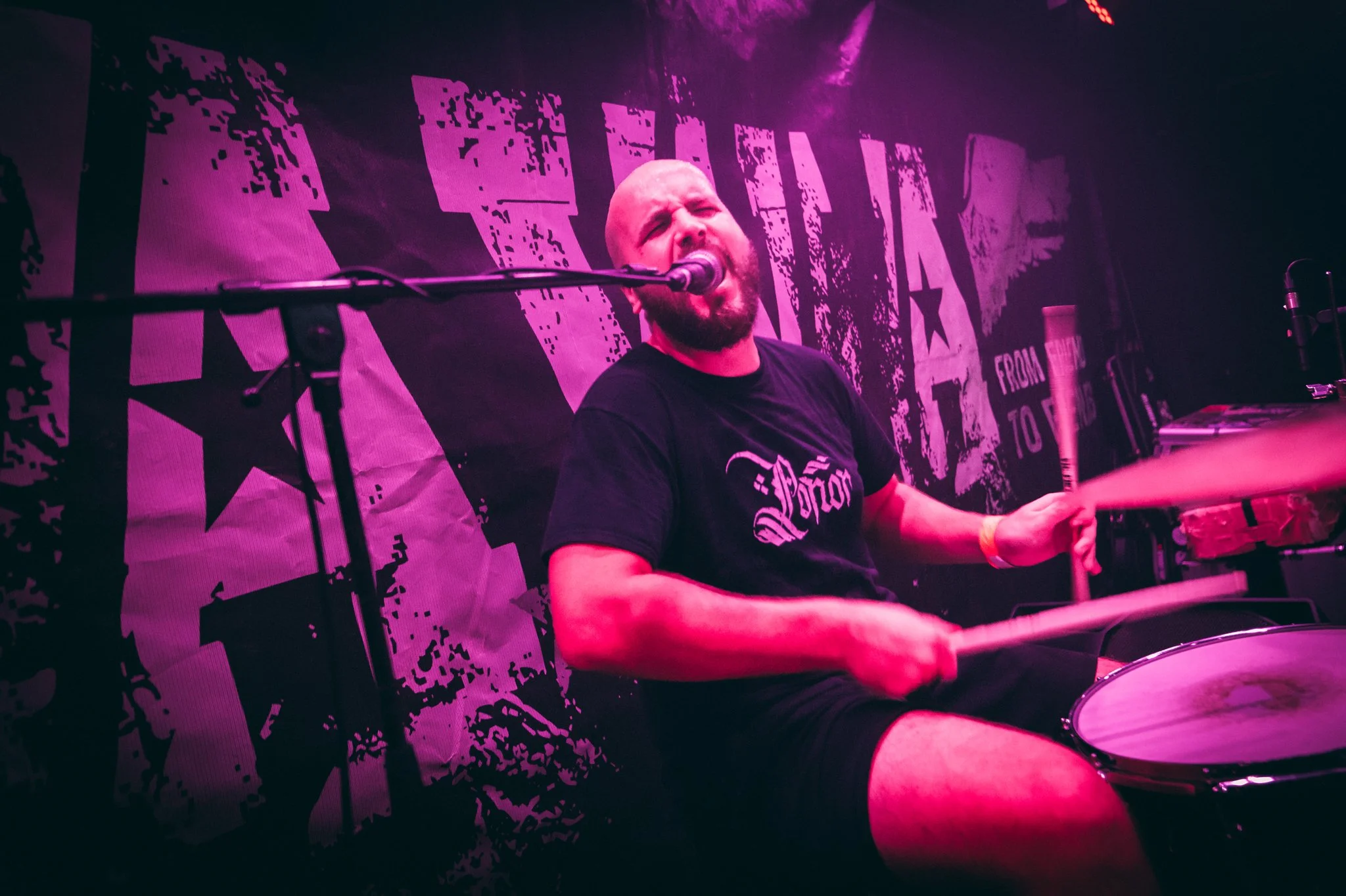Male drummer performing on stage, singing into a microphone, with purple lighting and a backdrop with bold graphics.