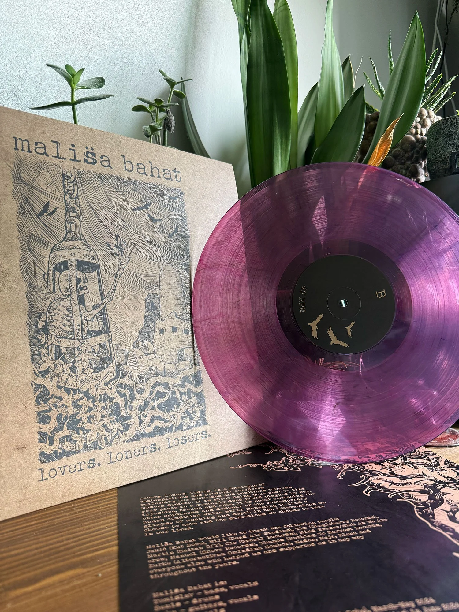 A record album in a purple transparent vinyl and a booklet featuring a black-and-white illustration of a skeleton with a chain and flying birds, and the title 'Lovers. Losers.'