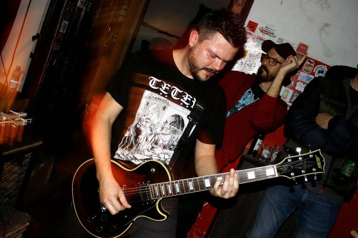 A man playing a black electric guitar with a label on the headstock, wearing a black T-shirt with a graphic design, in a room with various posters and stickers on the wall, and two other men standing nearby, one with glasses and a red shirt, and the 