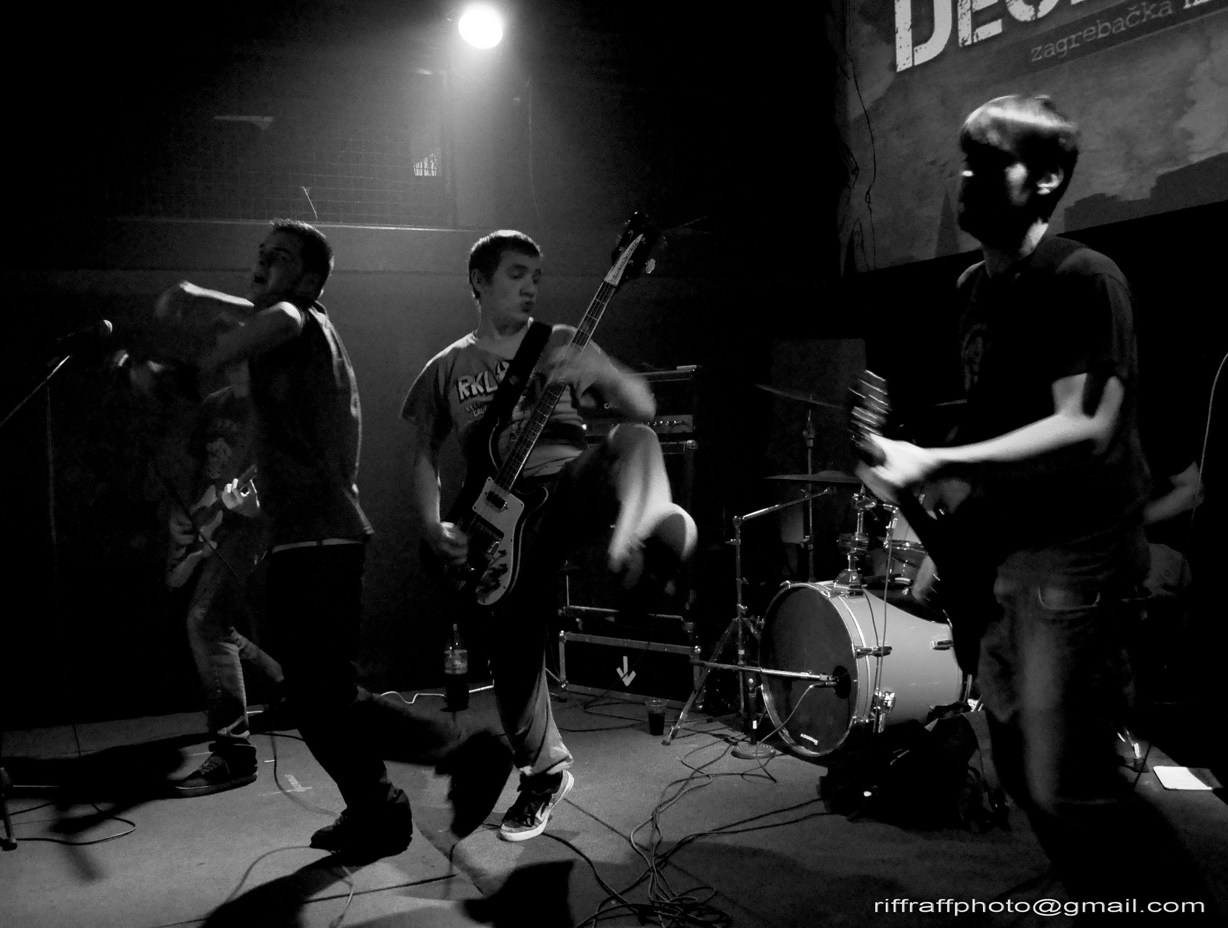 Four young men performing on stage, playing musical instruments including a guitar and drums, under stage lights.