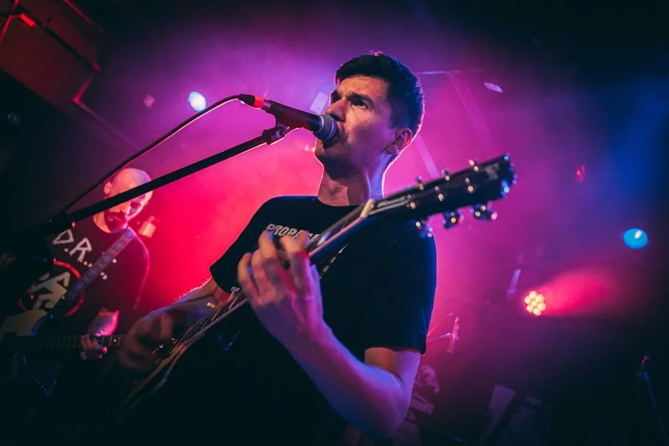 A male musician playing an acoustic guitar and singing into a microphone on stage with colorful stage lights.