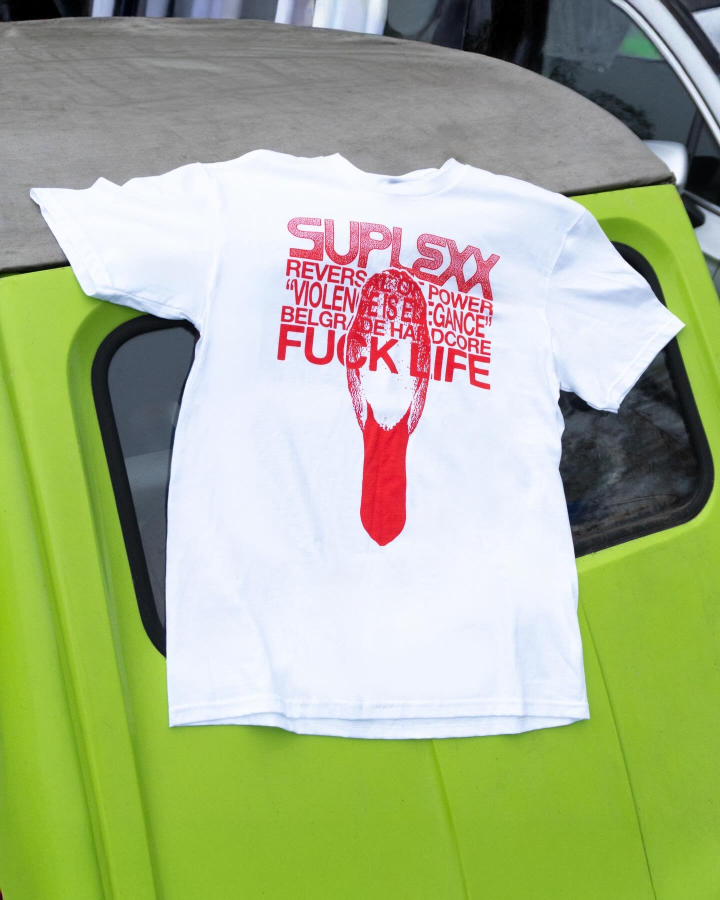 A white T-shirt with red text and graphics is draped over a green vehicle. The shirt has large text that reads 'SUP' and features a graphic of a hand flipping a bird, with more text including words like 'REVERS,' 'VIOLENCE,' 'POWER,' and 'FUCK LIFE'.