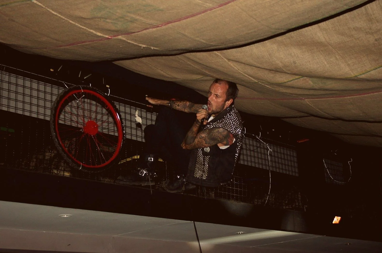 A male performer with tattoos singing into a microphone on a stage with a raised platform. The stage backdrop includes a bicycle wheel hanging on a wire grid.