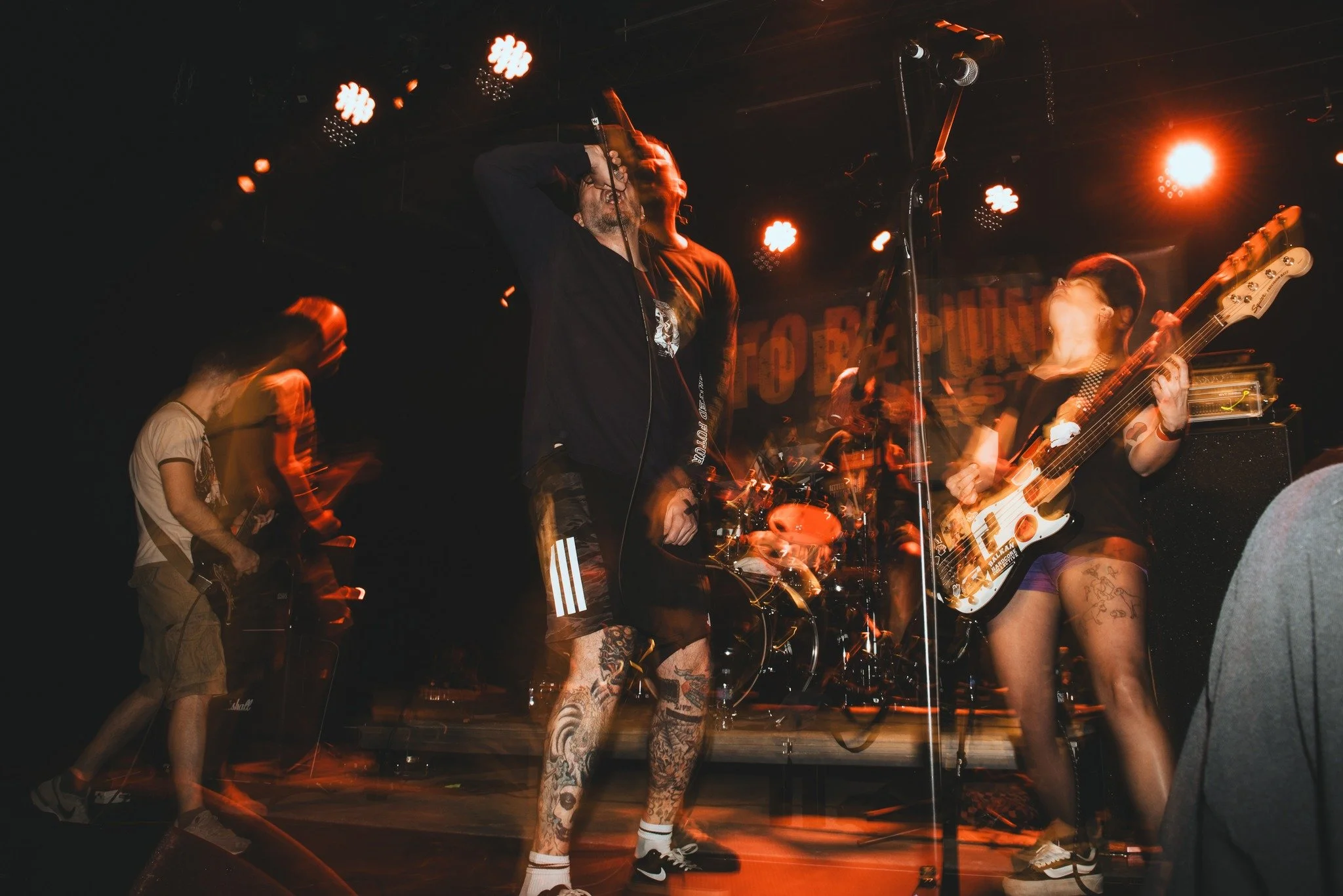 A band performing on stage at a concert, featuring a lead singer, a guitarist, and a bassist, with stage lights and musical equipment.