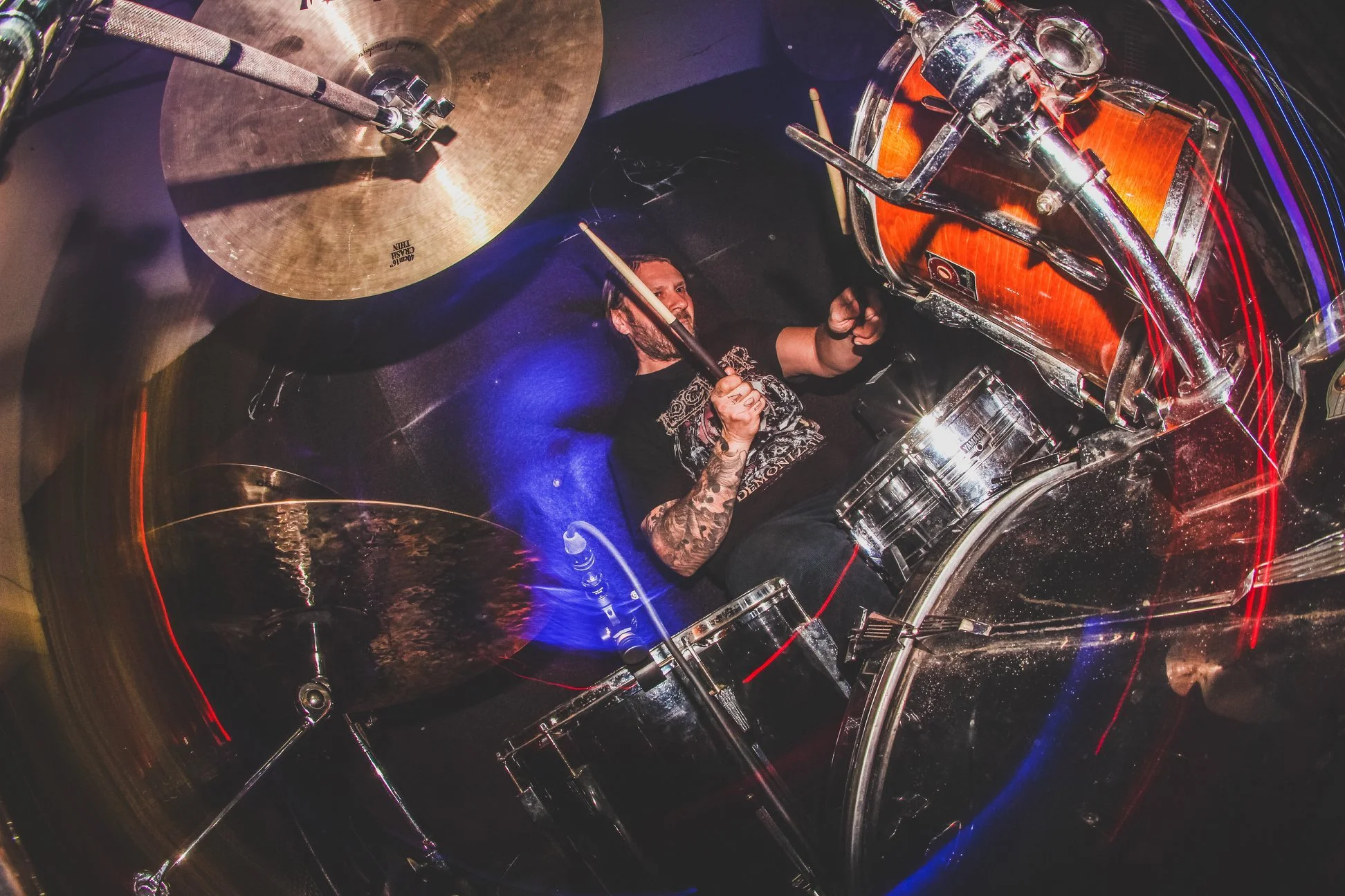 A drummer with tattoos playing drums inside a transparent drum set with colorful LED lights.