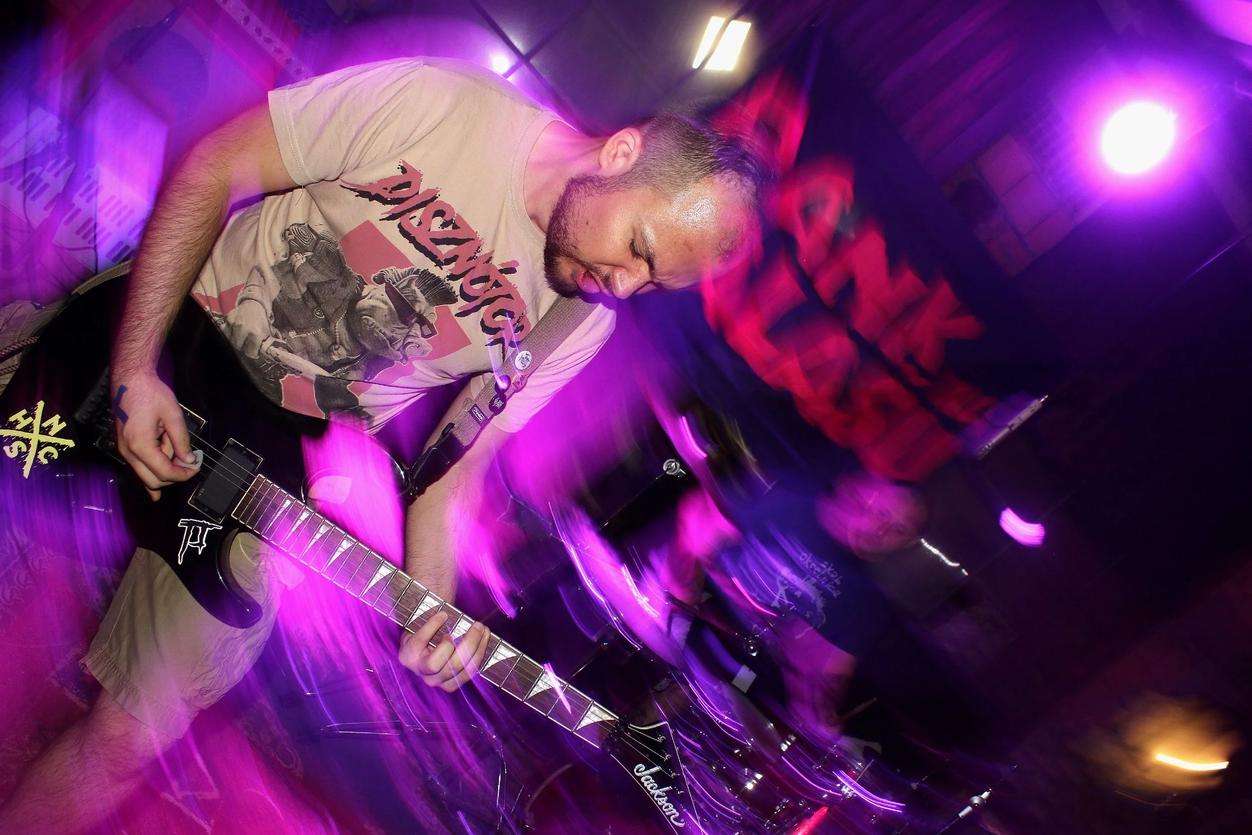 A man playing an electric guitar under vibrant purple and pink lighting at a music event or club.