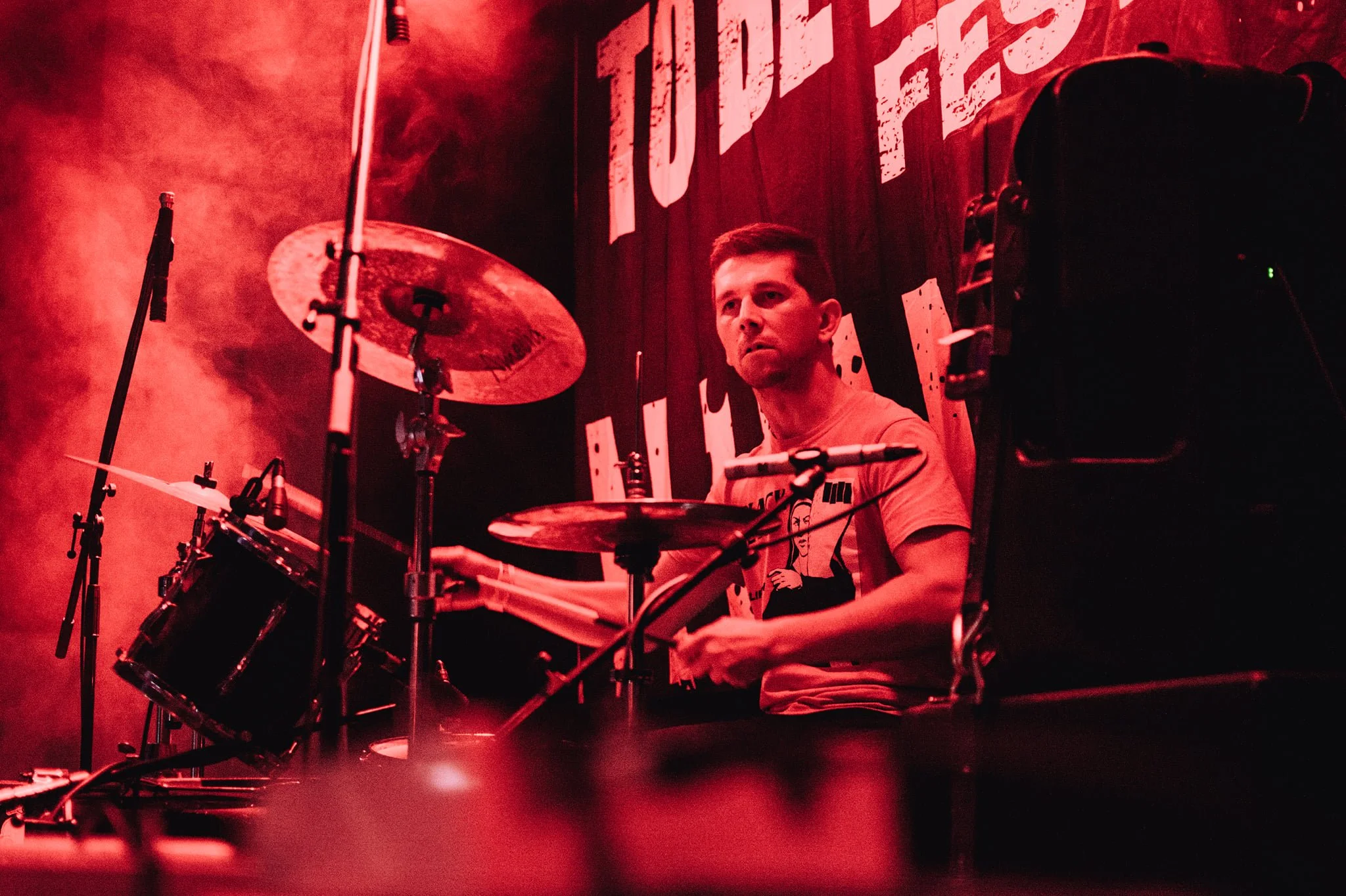 A young man playing the drums in a dimly lit, red-hued room, with a large banner behind him that partially reads 'STUBB'.