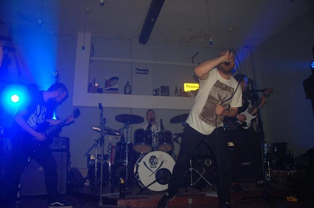 Band performing live music on stage with drums, guitar, bass, and vocalist, in dim lighting with blue and purple stage lights.