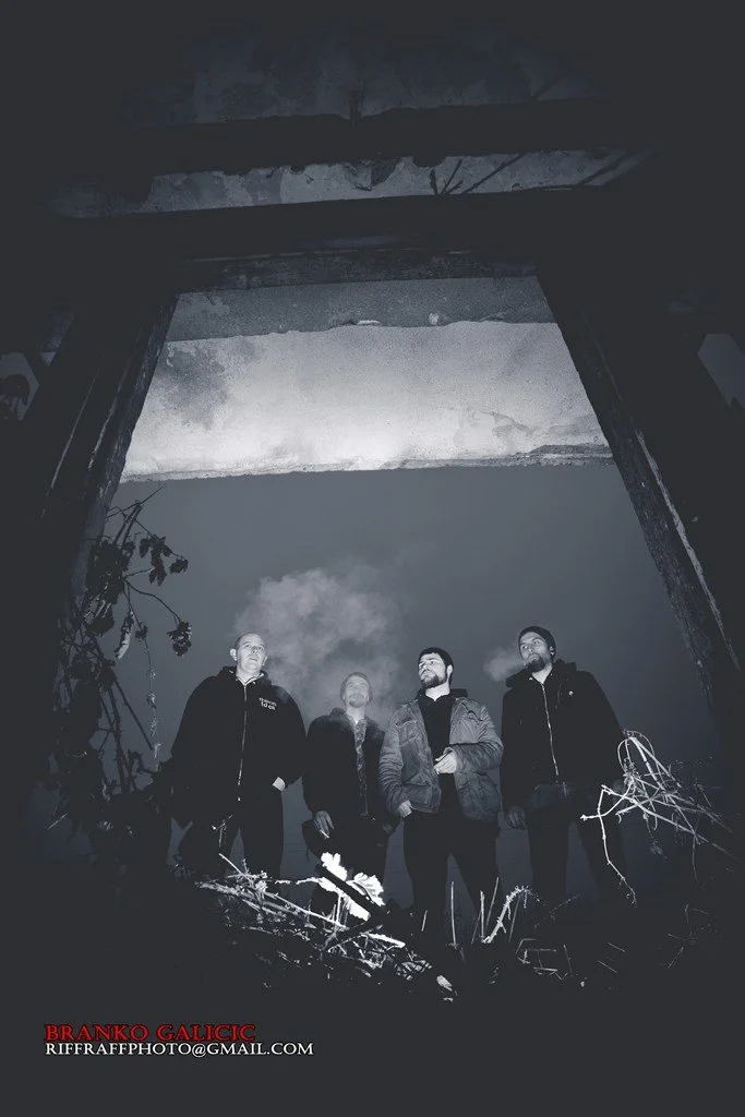 Four men standing outdoors at night, looking upward, with a cloudy sky and visible smoke or fog in the background. The photo is in black and white.
