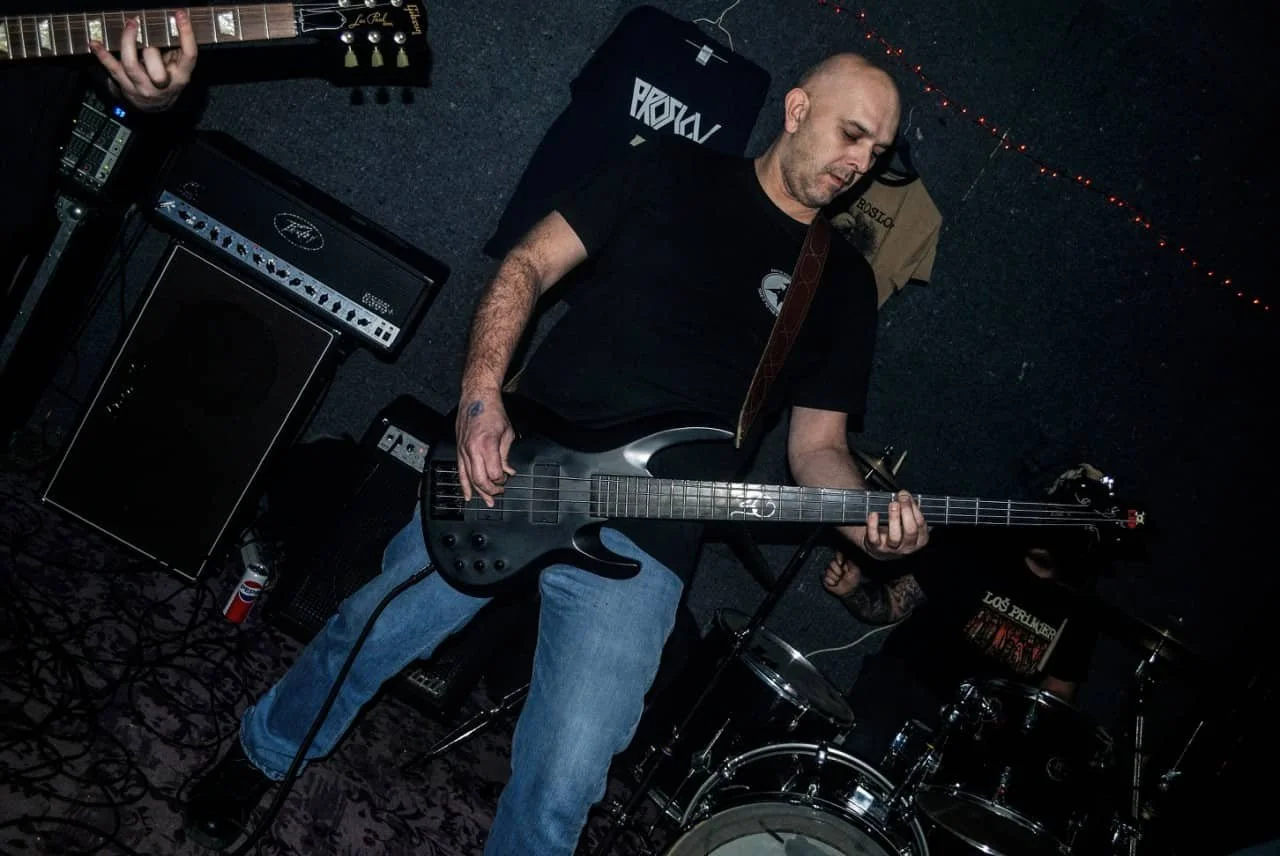A man playing a bass guitar in a music venue, with a drummer in the background and various musical equipment around him.