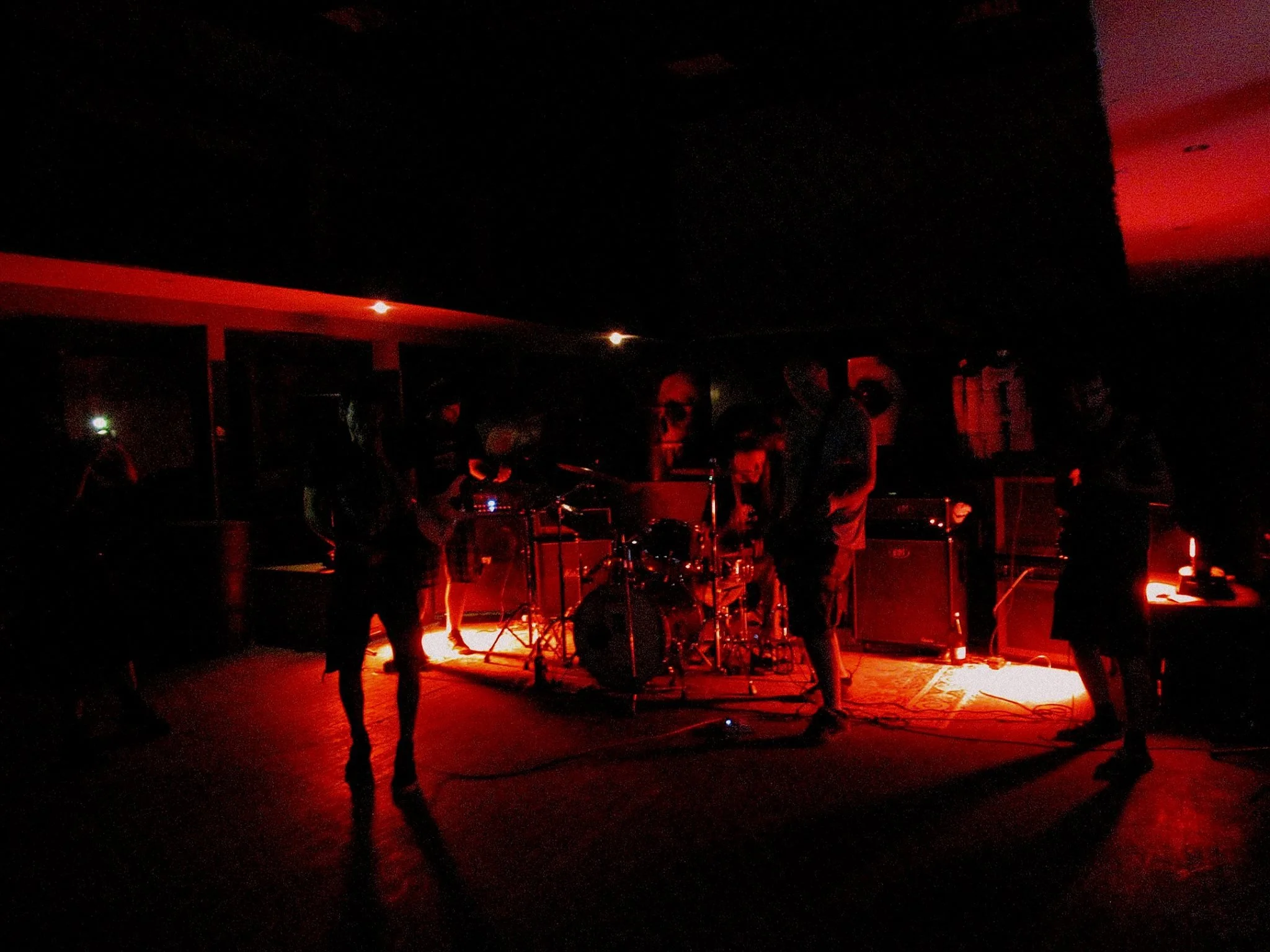 A dark indoor scene with a small band performing on stage illuminated by red and orange lighting, with several musicians and musical equipment visible.