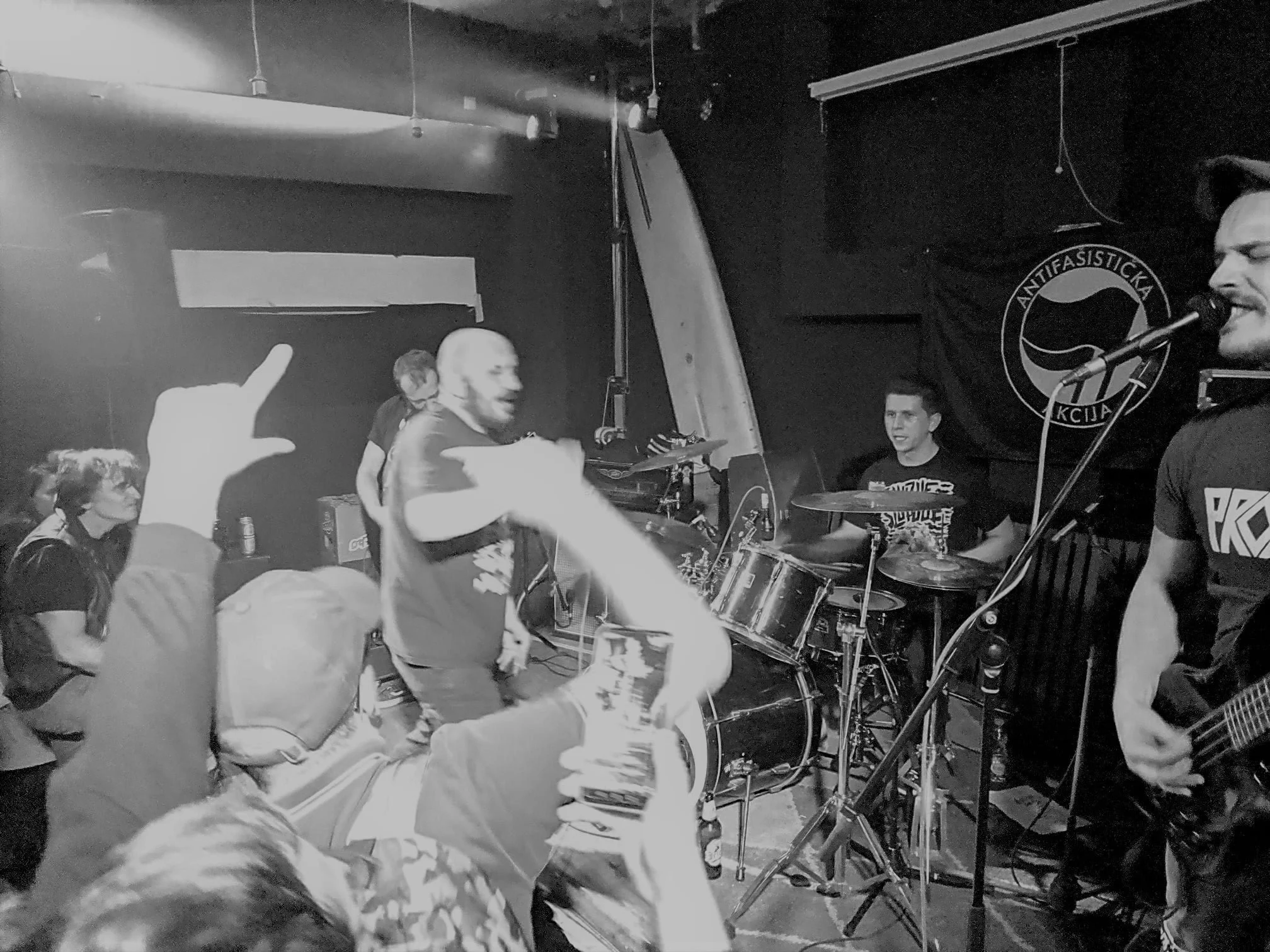 A black-and-white photo of a live band performance at an indoor venue. The image shows multiple band members playing instruments and singing, with a backdrop featuring a circular logo that says 'ANTIFASISTICKA.' The audience is visible with some peop