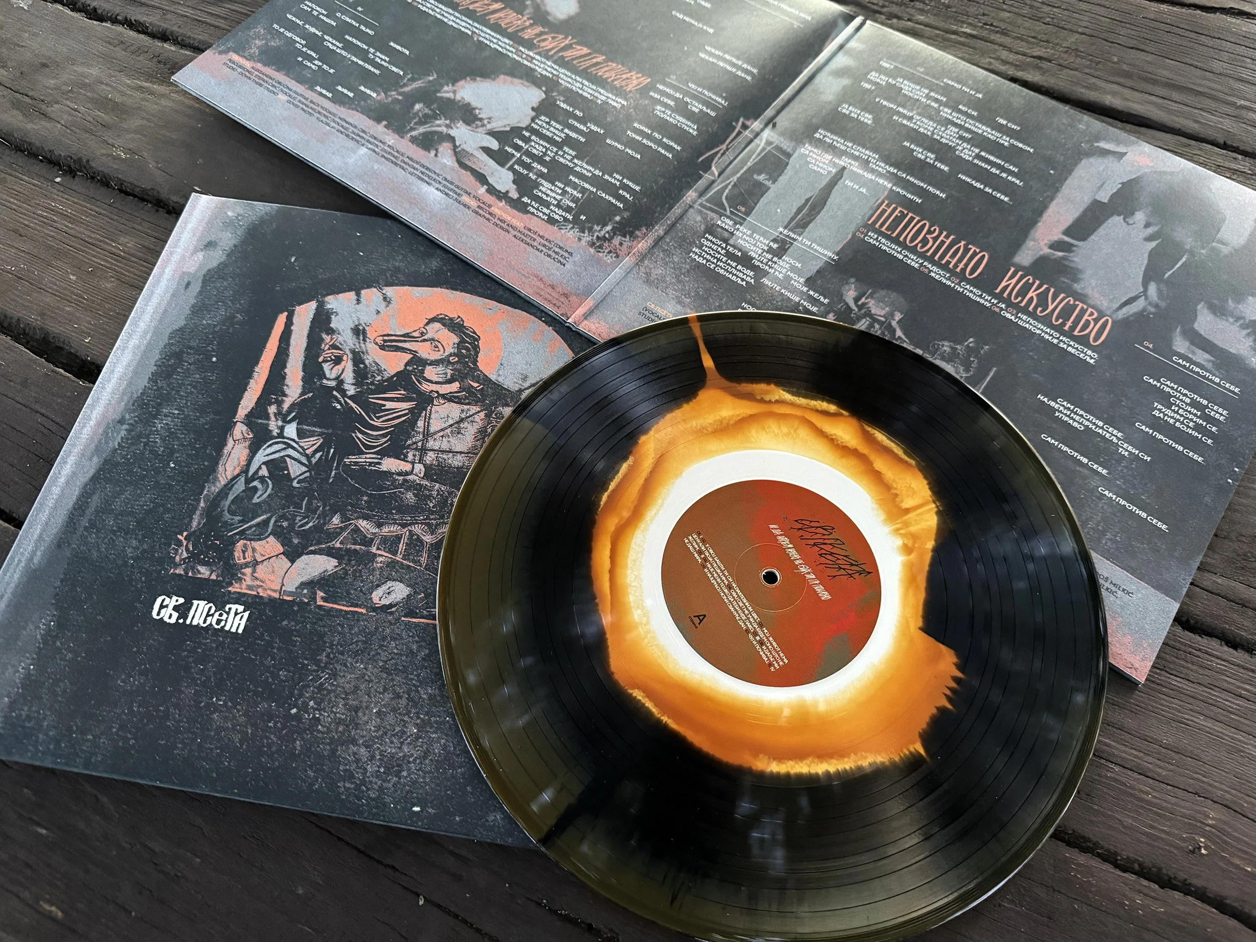 Photo of a vinyl record with an orange and yellow center label, resting partially on an open booklet on a wooden surface. The booklet contain text and illustrations, with one prominent illustration of a person wearing a beak mask. SVETA PSETA