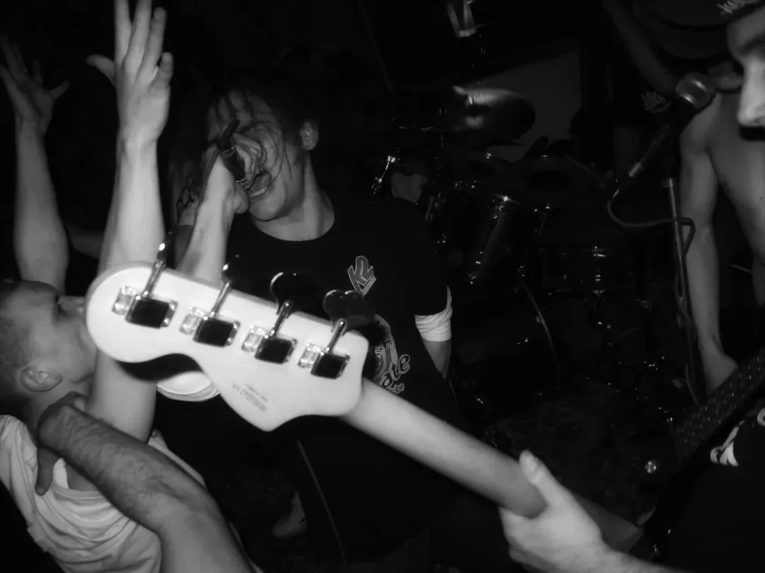 Black and white photo of a live music scene with people close together. A person in the center is singing into a microphone, with long hair covering part of their face. A person on the right is playing a guitar. A drum set is visible in the backgroun