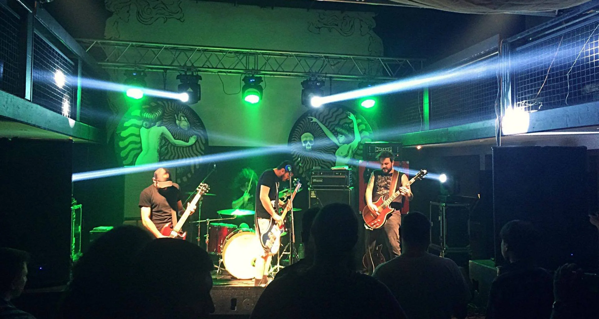A live band performs on stage with green and white lighting, logo art on the wall featuring a mermaid and skull, audience silhouettes in foreground. Band name: 1000mods