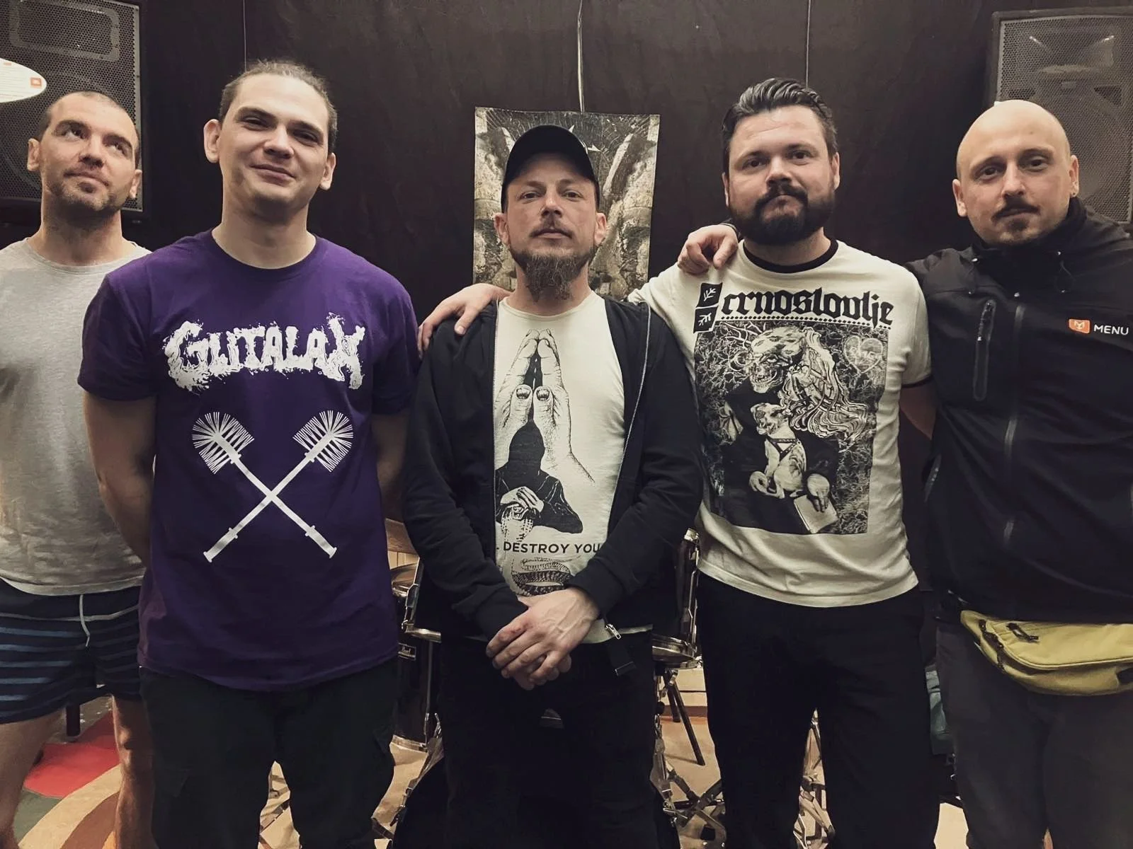 Group of five men standing together in a room with black walls, some wearing band t-shirts, and one with yellow fanny pack, posing for a photo.