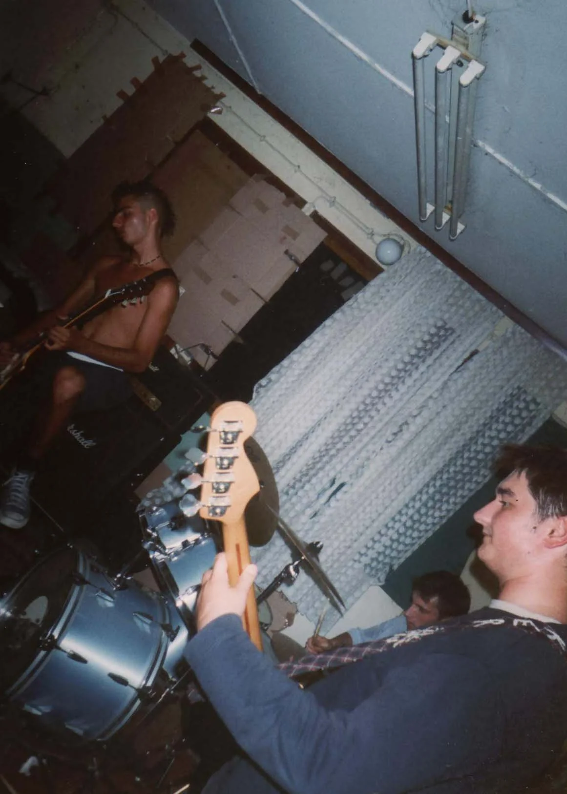 A group of young musicians playing instruments in a basement or rehearsal space, including a shirtless guitarist, a drummer, and a bassist in the foreground, with a curtain and wall pipes in the background.