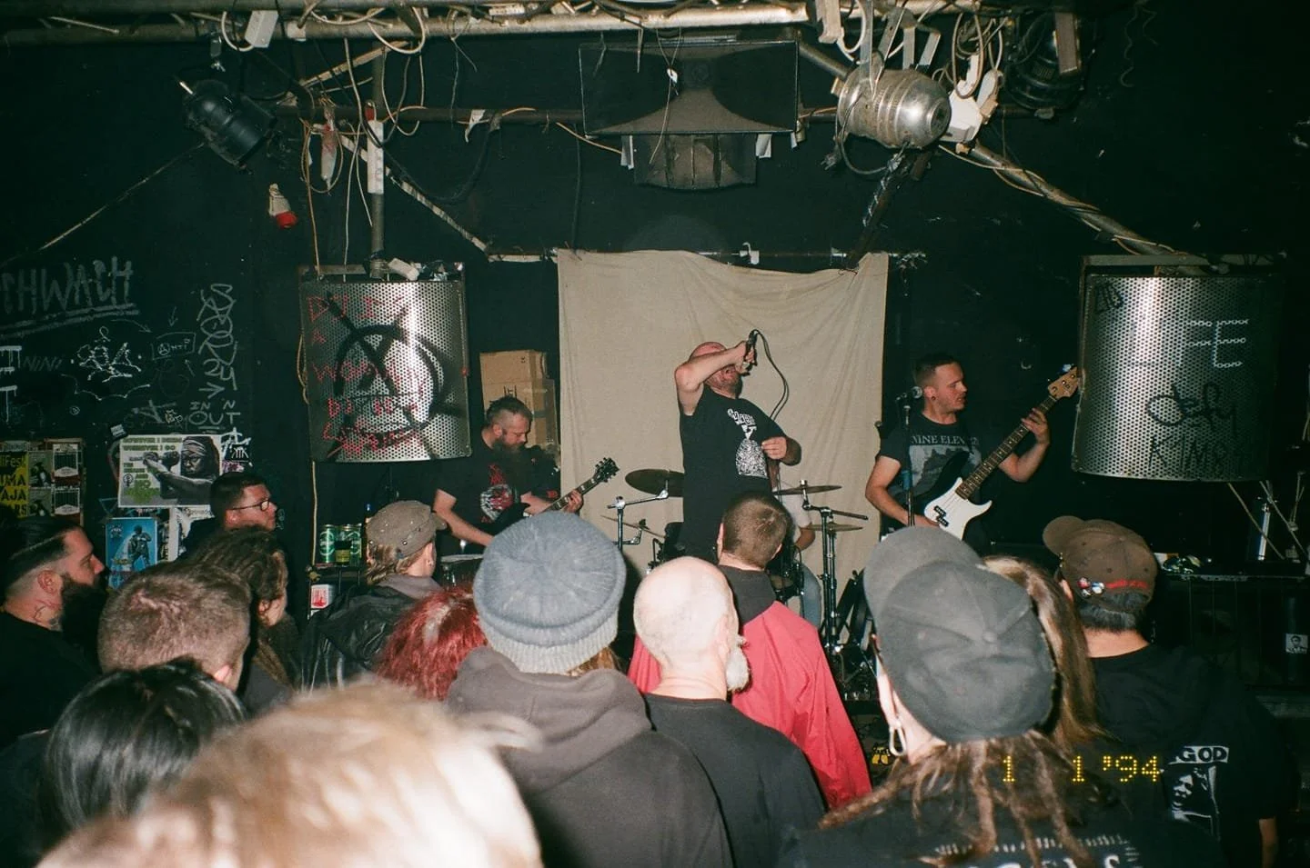 A live band performing on stage in a small venue with an audience watching, with graffiti and posters on the black walls.
